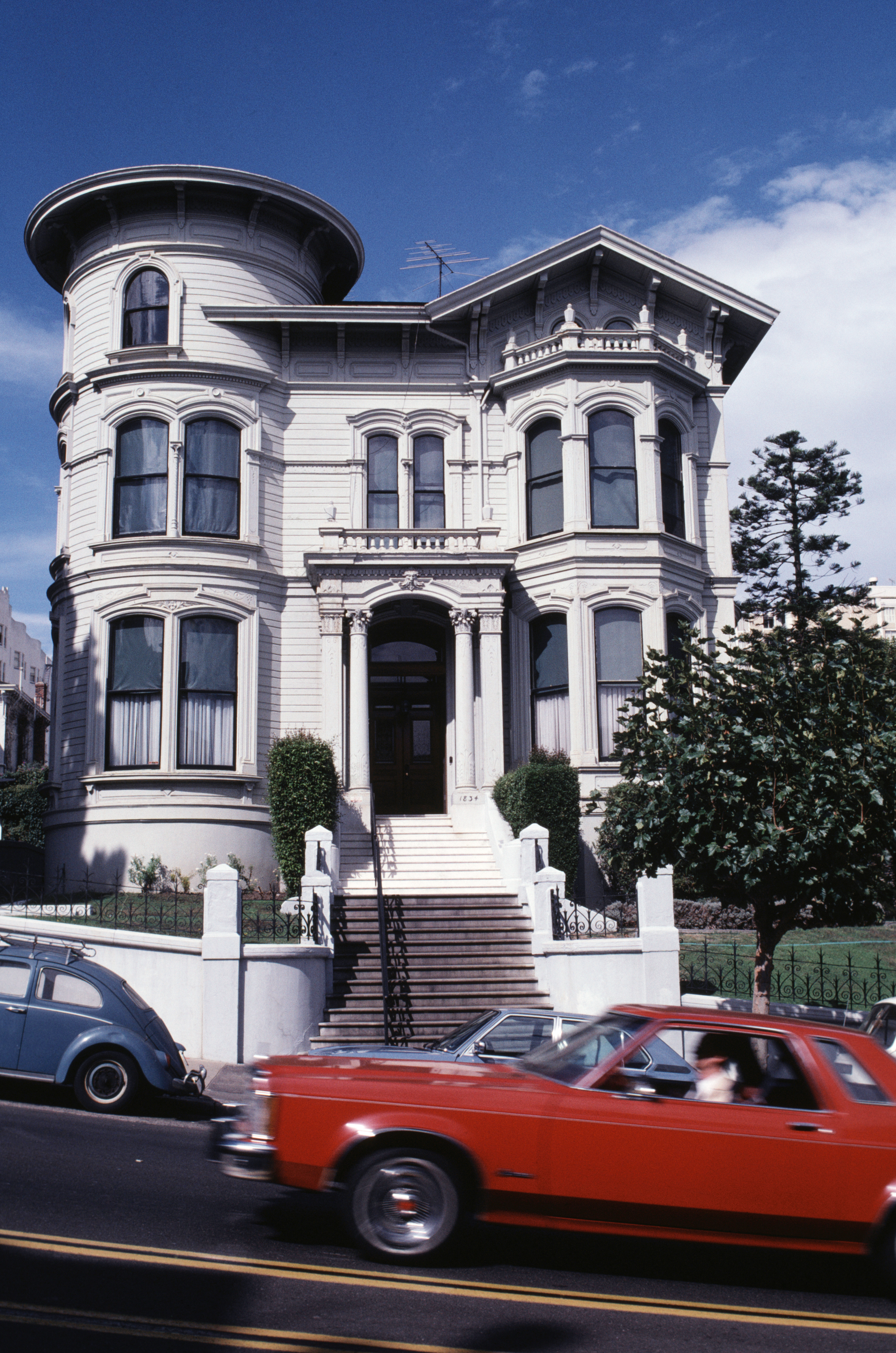 A historic, Victorian-style house with large bay windows and a rounded tower stands along a street. A red car passes by in the foreground