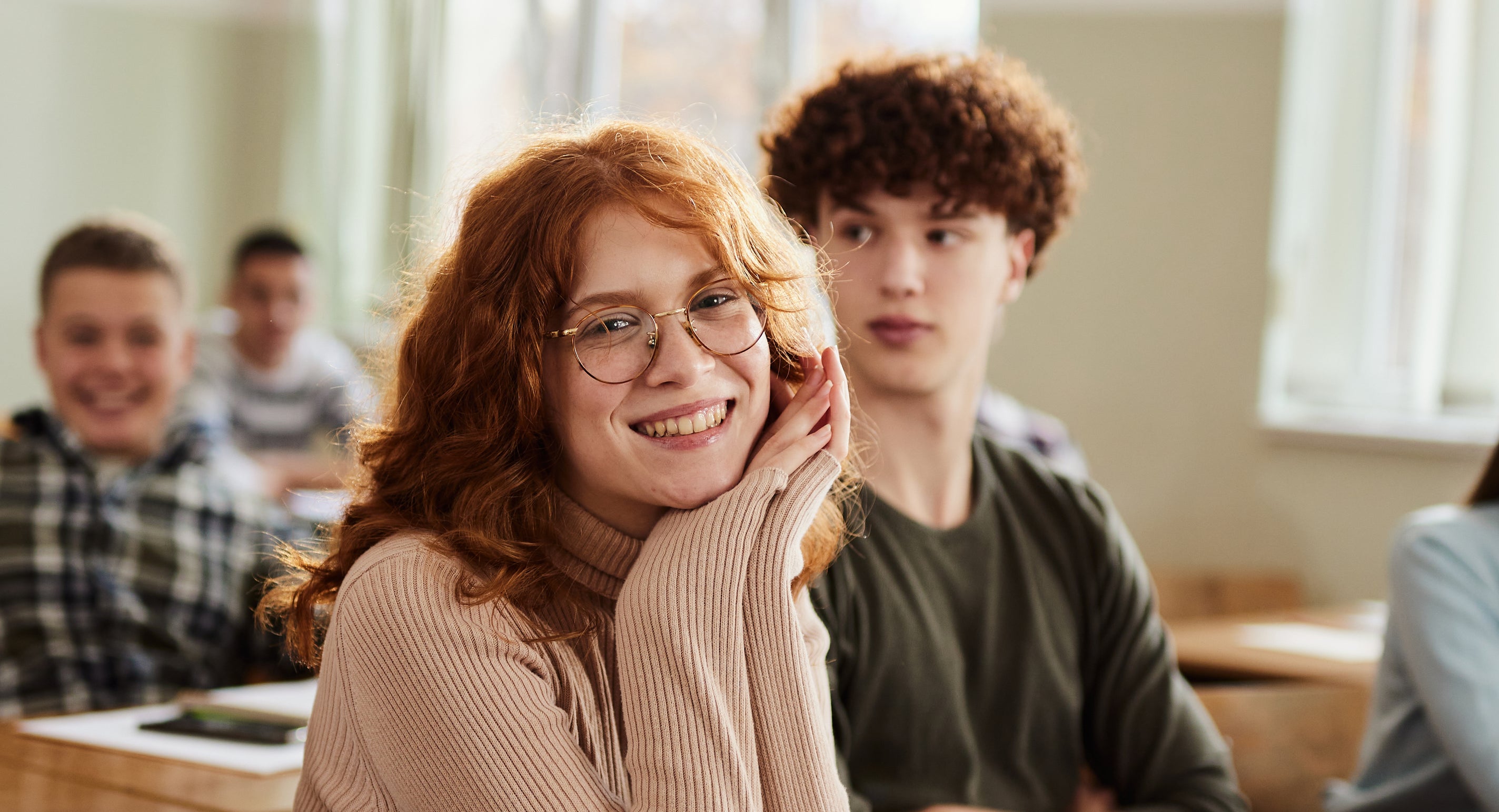 A group of students, including a smiling girl with red hair and a boy with curly hair, sit in a classroom at their desks with notebooks in front of them