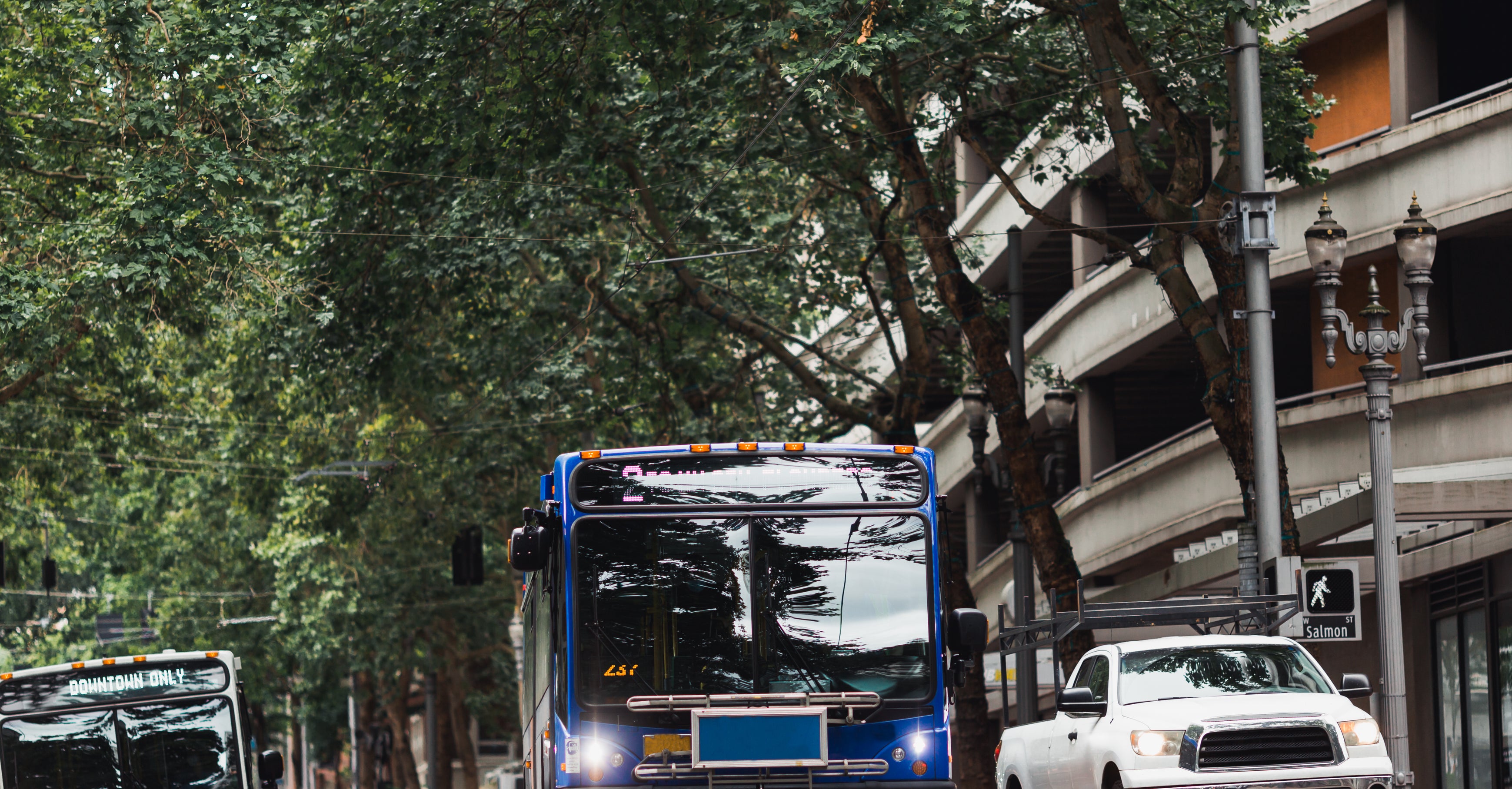 Two city buses and a white truck on a tree-lined urban street near a multi-story parking garage