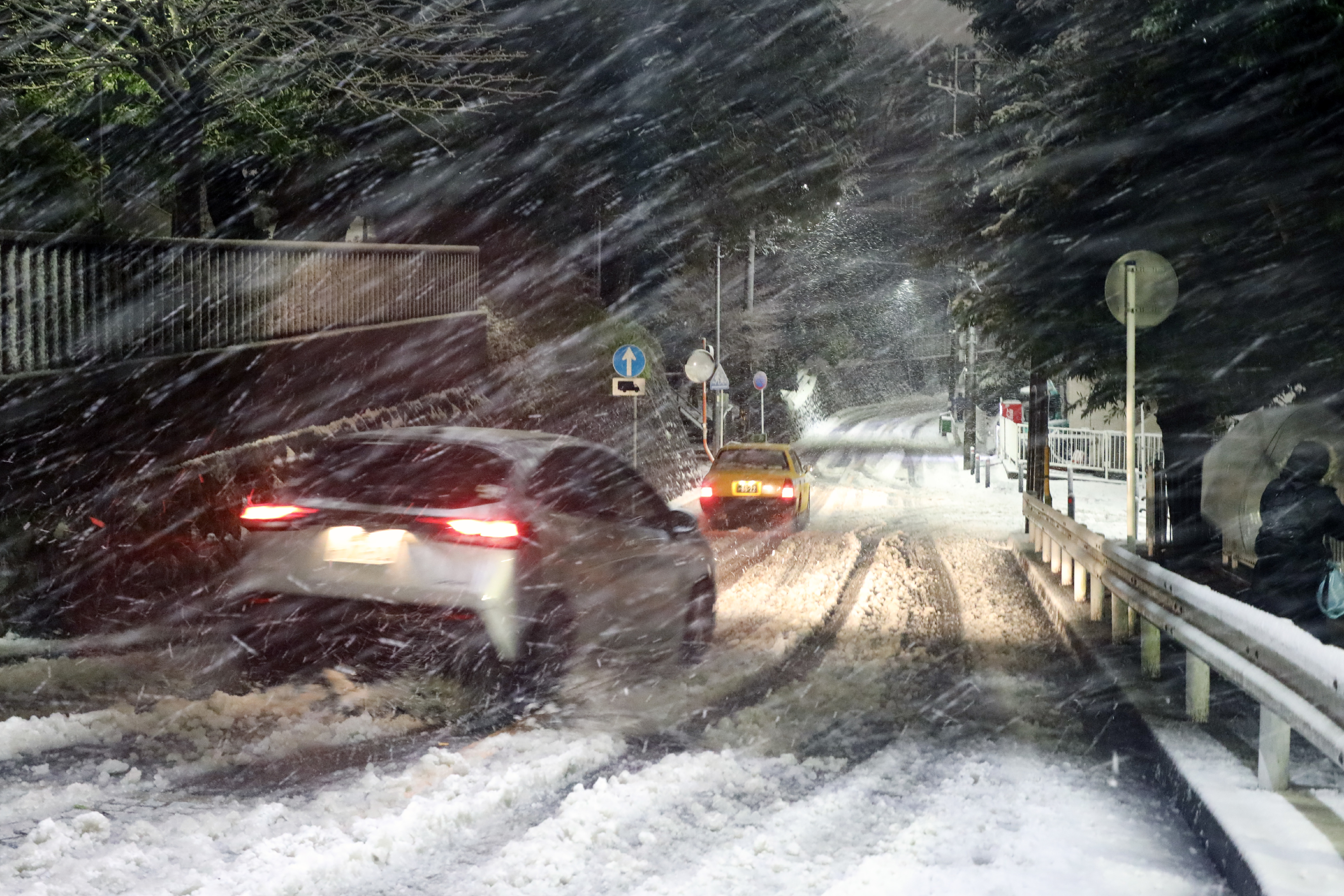 Cars navigate a snowy, stormy road with decreased visibility, heavy snow falling, and snow-covered surroundings