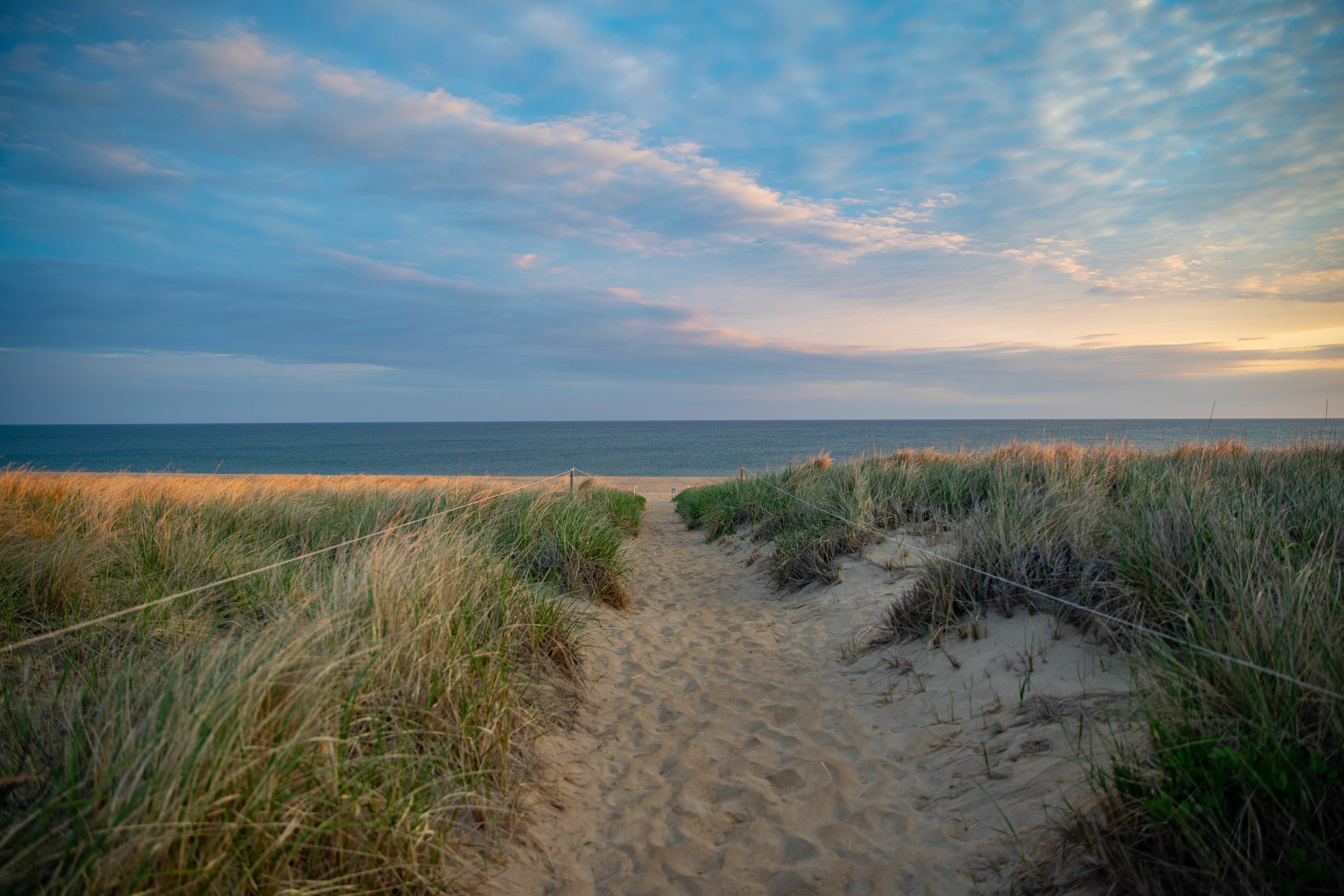 Path through sand dunes leading to the ocean under a partly cloudy sky at sunset