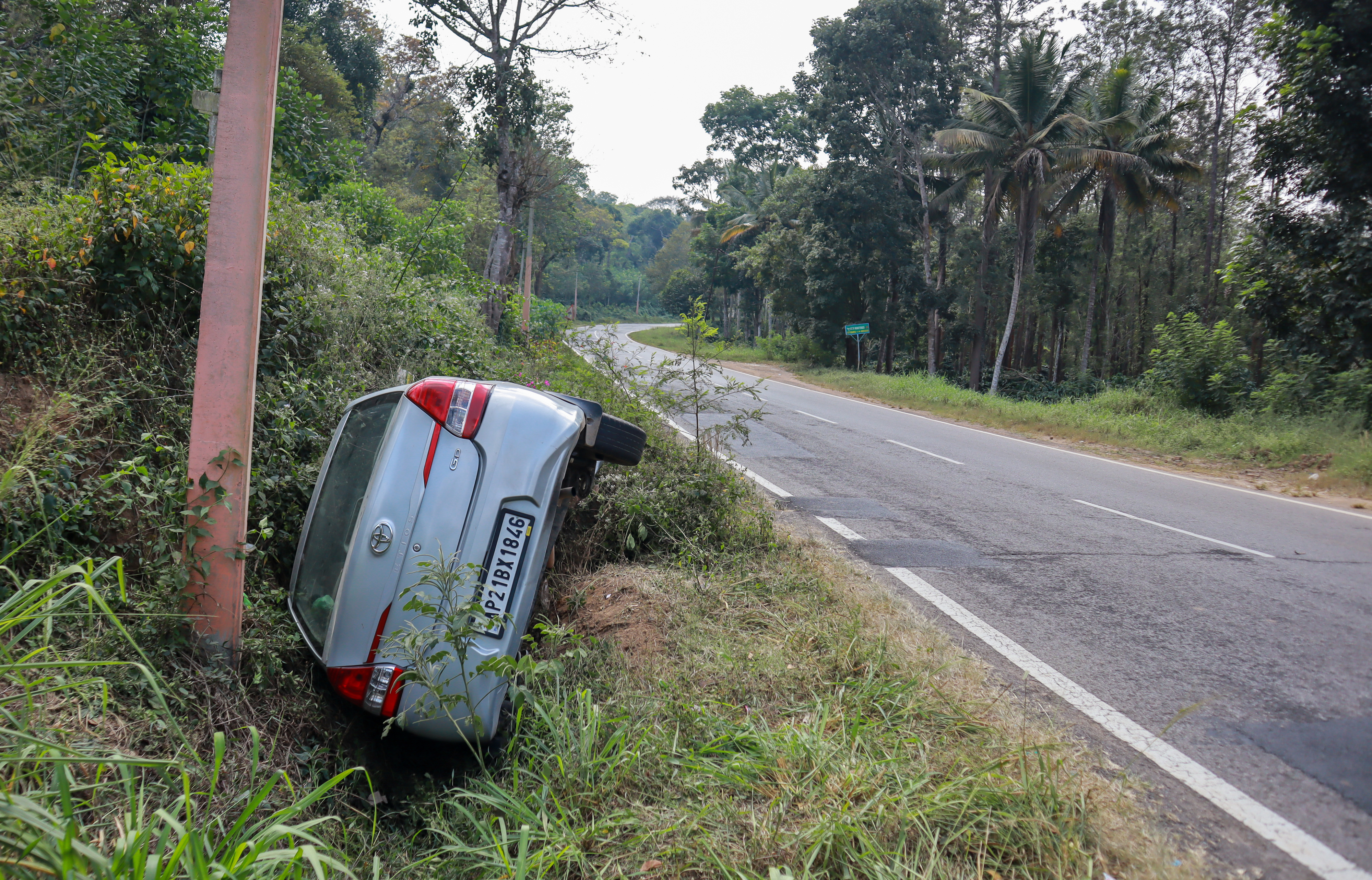 A silver car is overturned in a ditch beside a rural road, with its rear end up against a utility pole. The area is surrounded by trees and greenery
