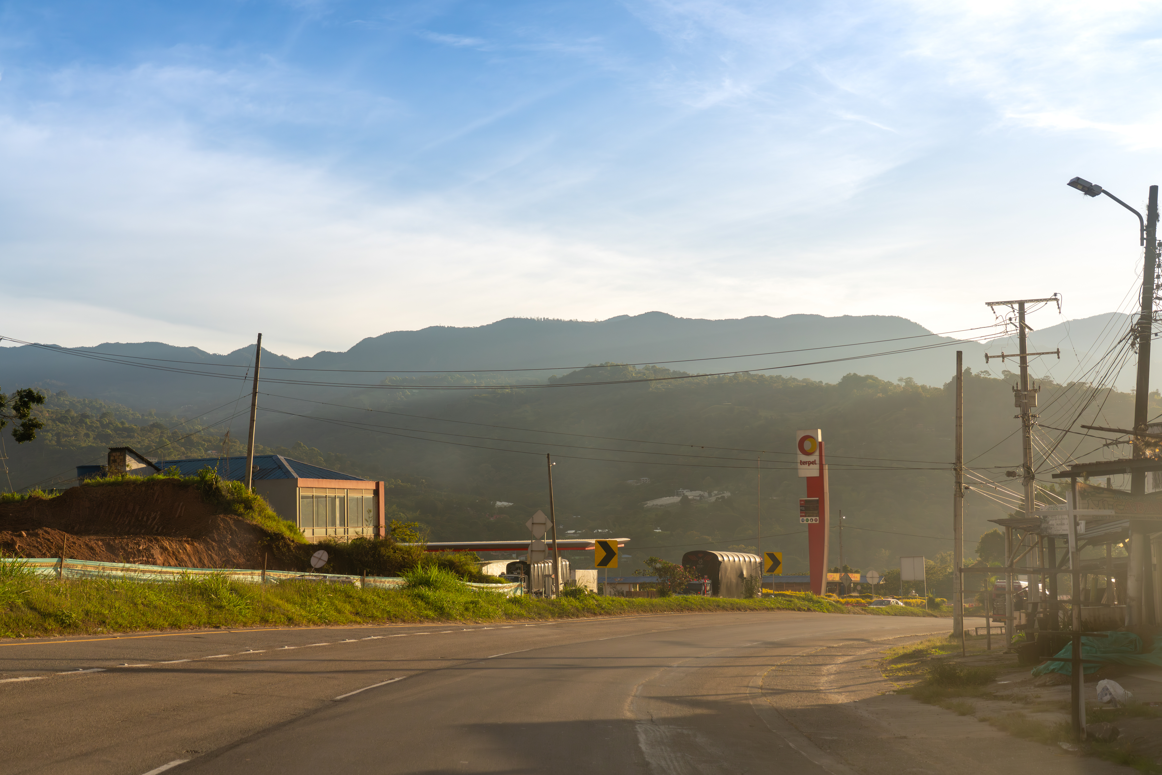 A scenic view of a rural road approaching a gas station, with mountains and a clear sky in the background.