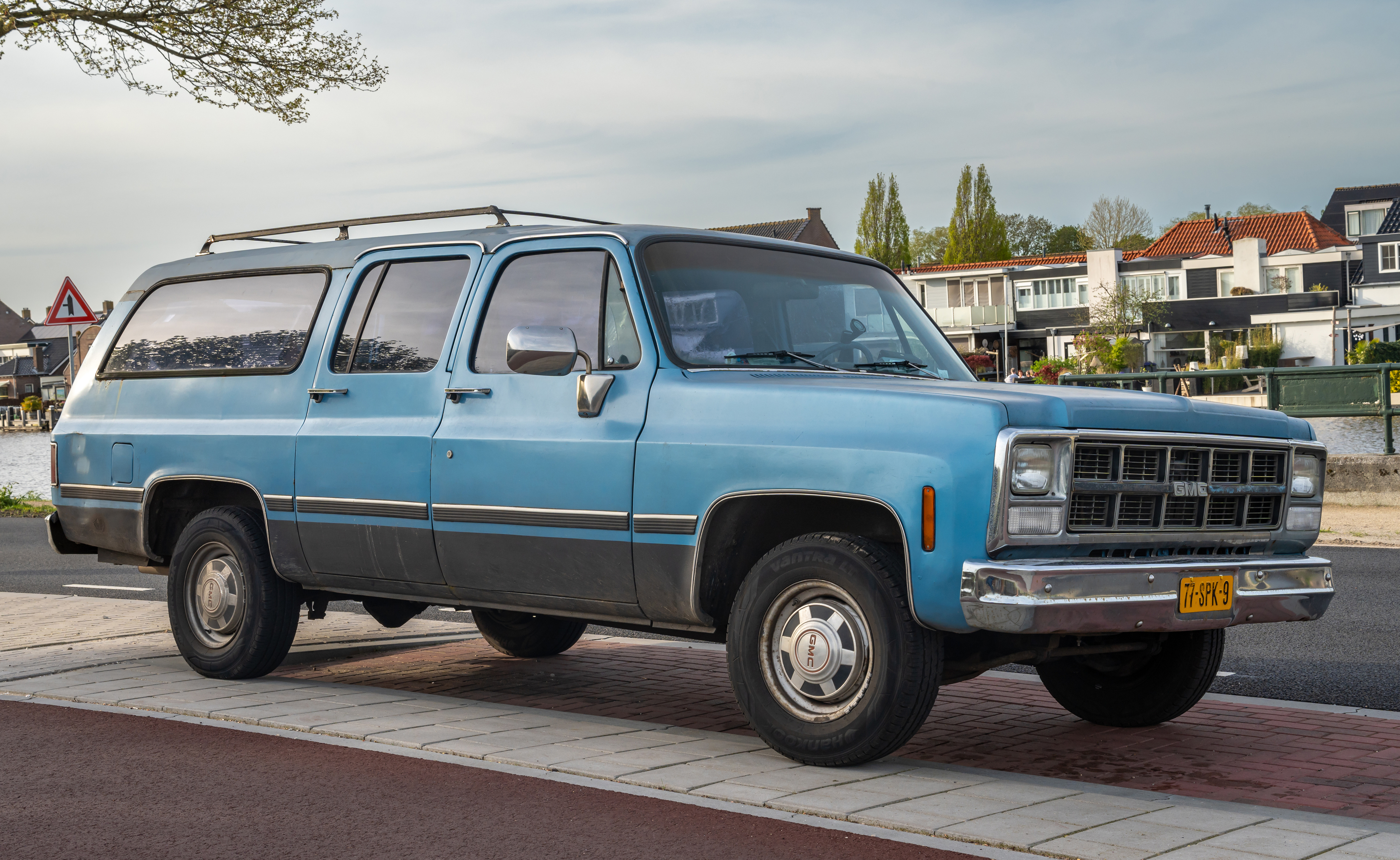 A vintage, blue Chevrolet Suburban parked on a sidewalk in a suburban area with houses and trees in the background