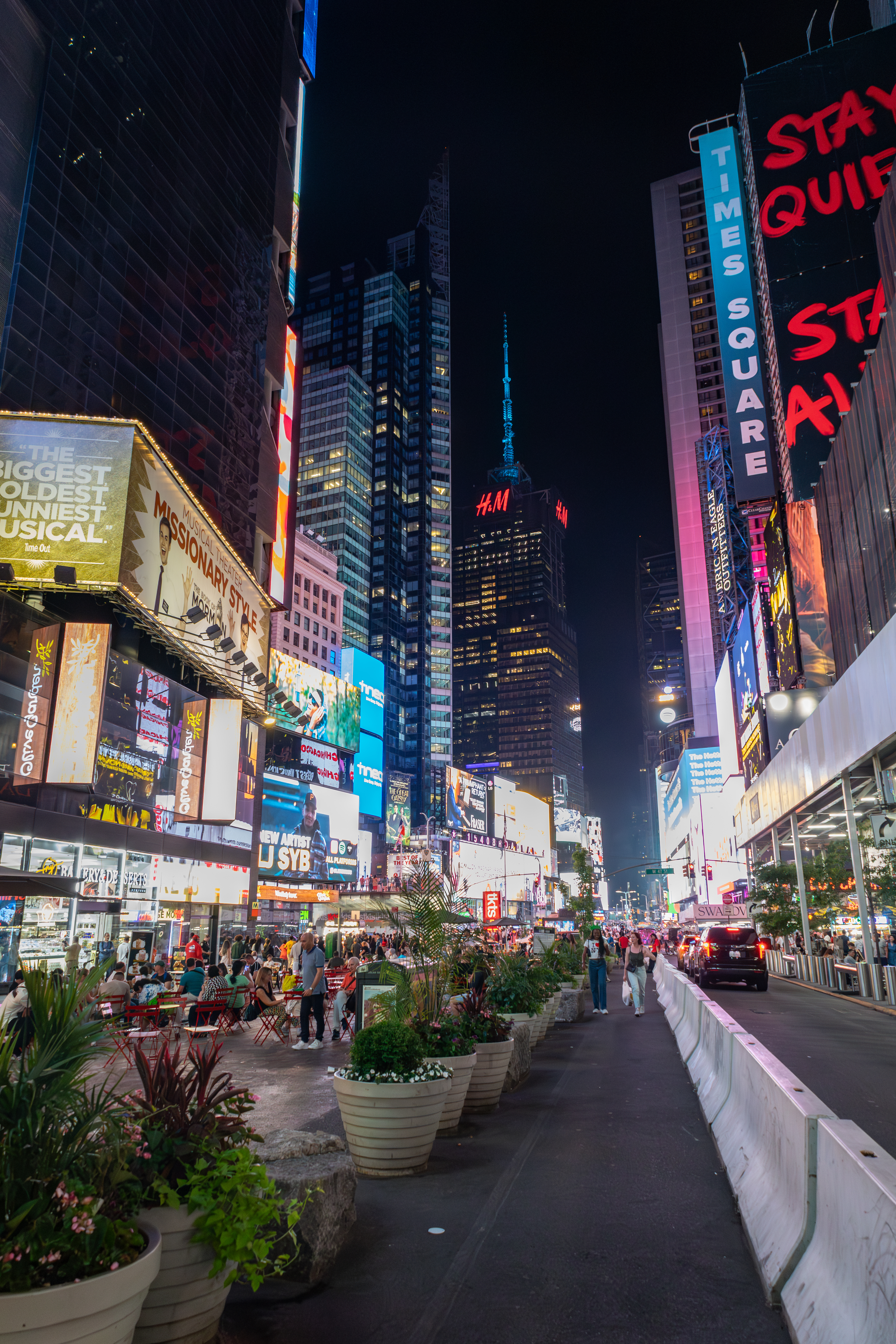 Times Square at night, bustling with pedestrian activity, bright electronic billboards, and s for shows and brands like H&amp;amp;M and Missionary Style