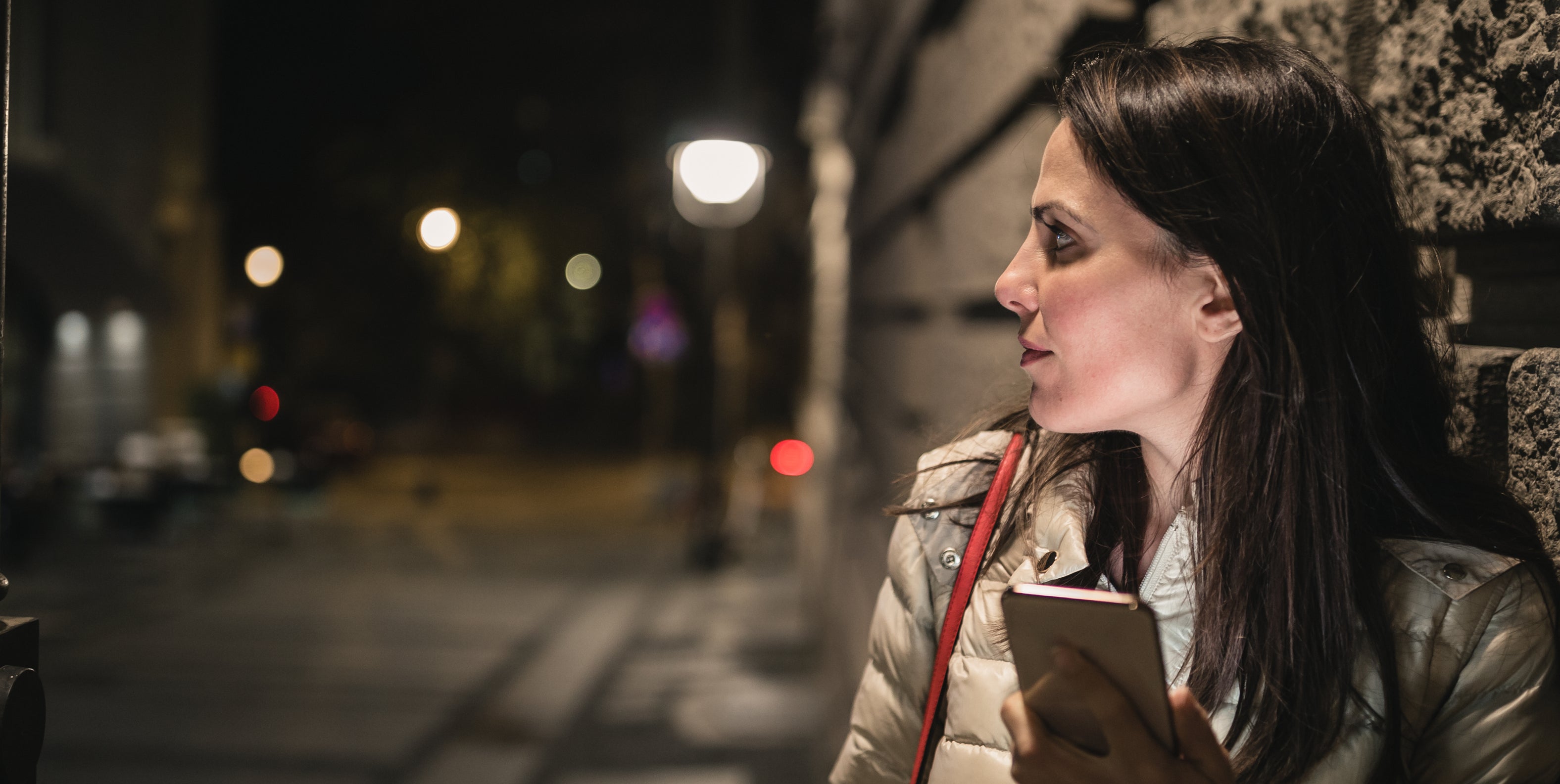 A woman in a puffy jacket holds a smartphone and leans against a stone wall on a city street at night, looking thoughtfully to the side