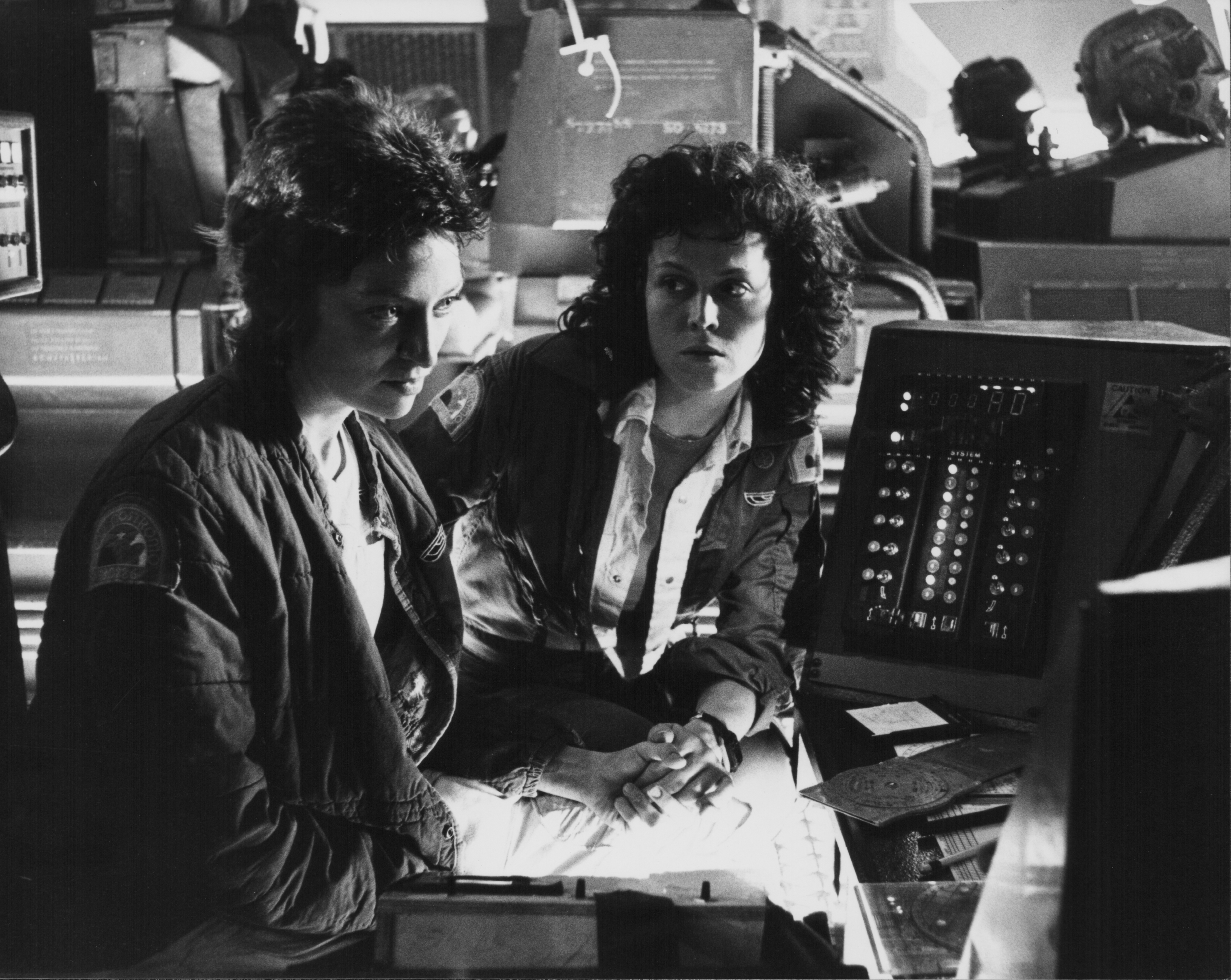 Sigourney Weaver and Veronica Cartwright in sci-fi film set, sitting beside control panels, wearing jackets, focused expressions