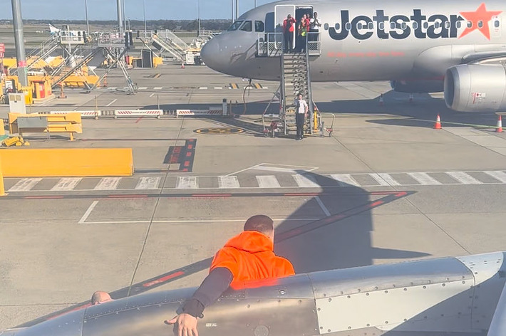 ジェットスターの飛行機の翼の上にオレンジのベストを着た整備員が乗っている様子。背景には搭乗中の乗客が見える。