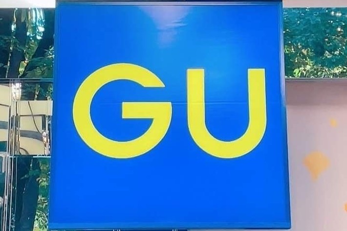 Sign in a store with large yellow letters &ldquo;GU&rdquo; on a blue background. The surrounding area features a reflective surface and some greenery