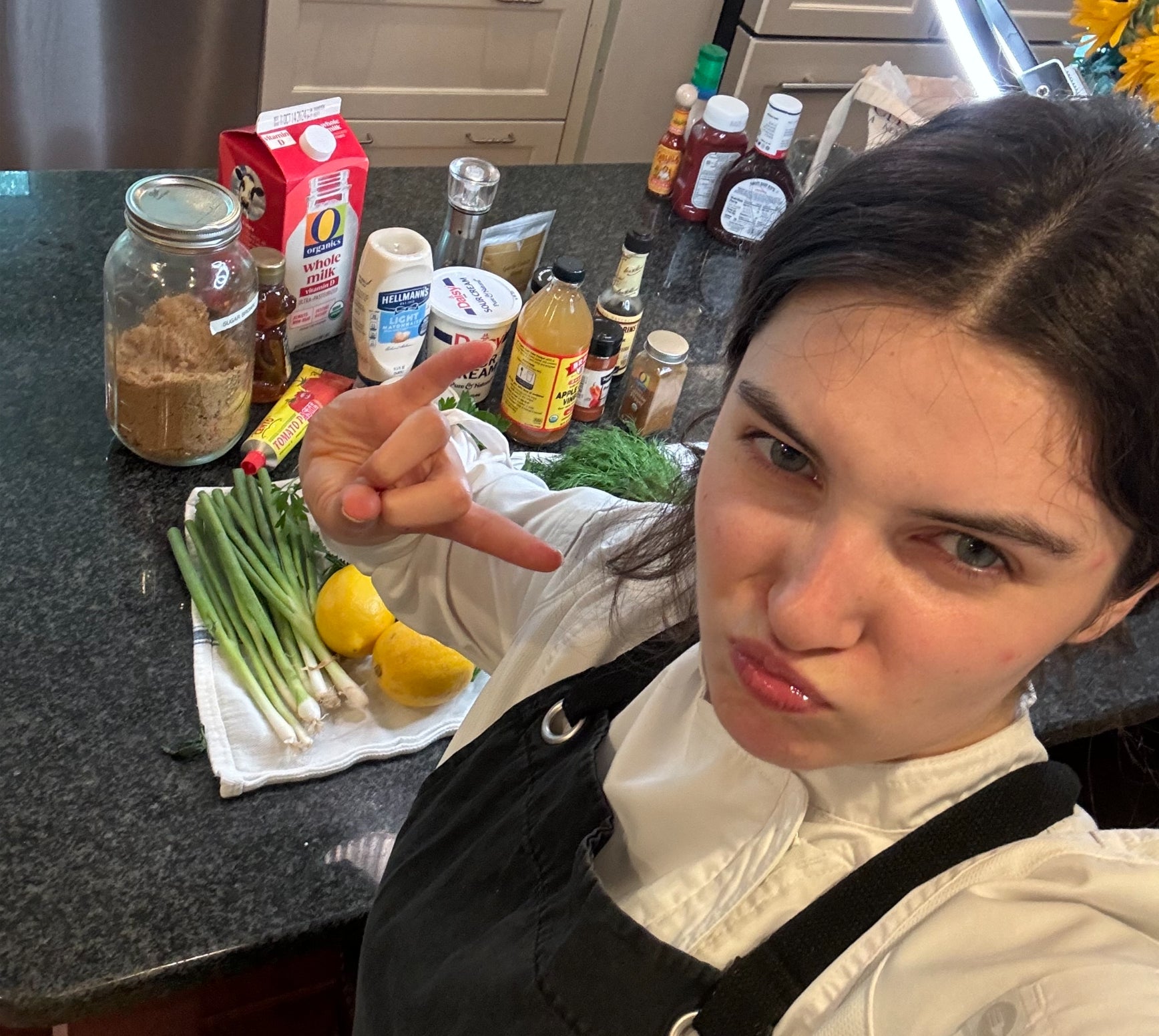 Person in a kitchen with various cooking ingredients on a counter, posing with a playful facial expression. Not a celebrity