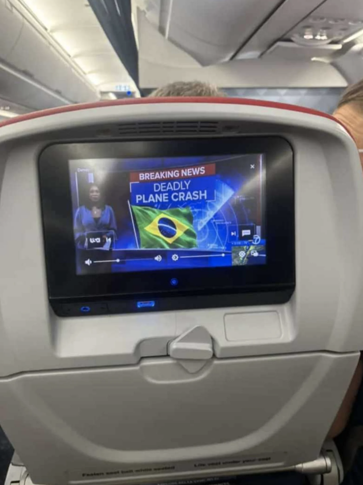 An airplane seatback screen displays breaking news about a deadly plane crash involving Brazil, with an anchorwoman reporting in the background