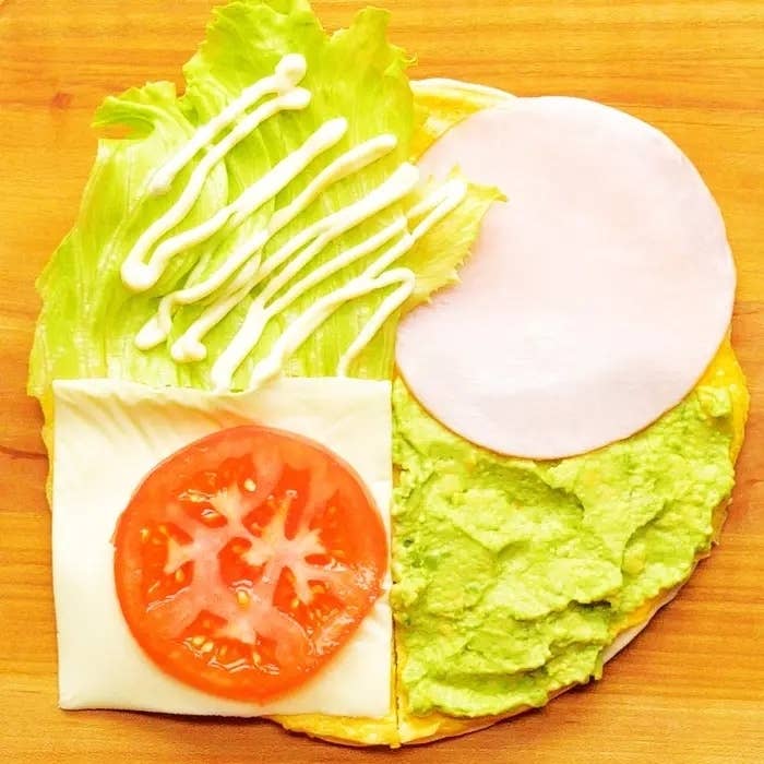 サンドイッチの具材が木のテーブルの上に並べられています。レタス、ハム、アボカドペースト、トマト、チーズがあります。