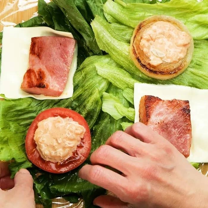 レタスにベーコン、チーズ、トマト、玉ねぎが置かれています。手が材料を包もうとしています。