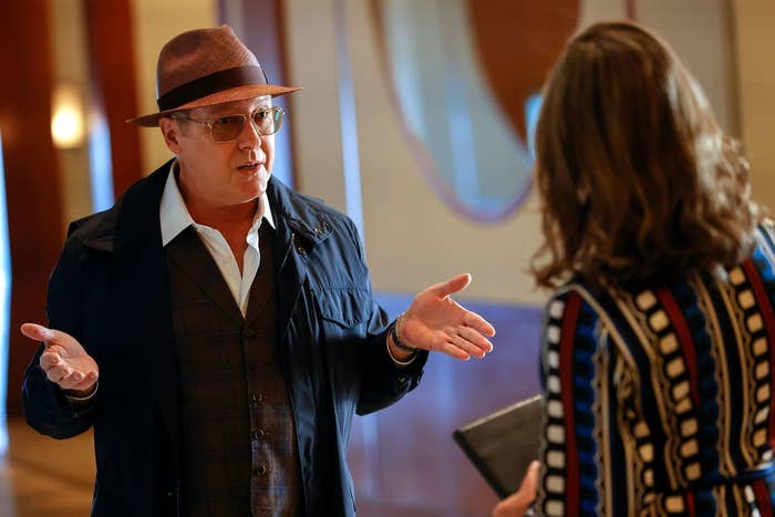 James Spader, wearing a hat and glasses, gestures with his hands while speaking to a woman in a patterned dress in a formal setting