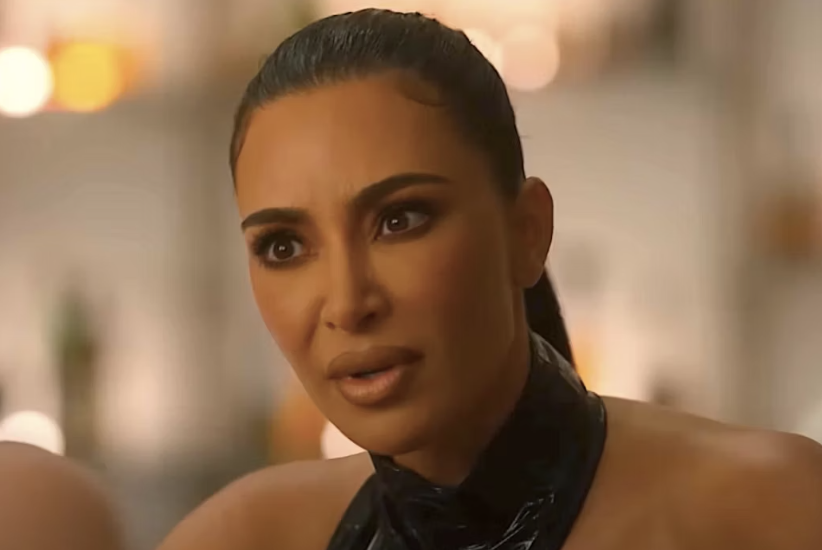 Kim Kardashian in an indoors setting, wearing a sleek, high-neck outfit, looking intently ahead with a concerned expression