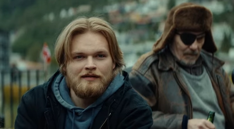 Two men outdoors; one with blonde hair and a beard wearing casual attire, another older man with an eyepatch and fur hat in the background