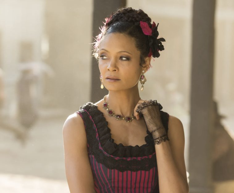 Thandie Newton wearing a Victorian-style dress with lace details, flower hair accessory, and jewelry. She gazes into the distance in an old-town setting