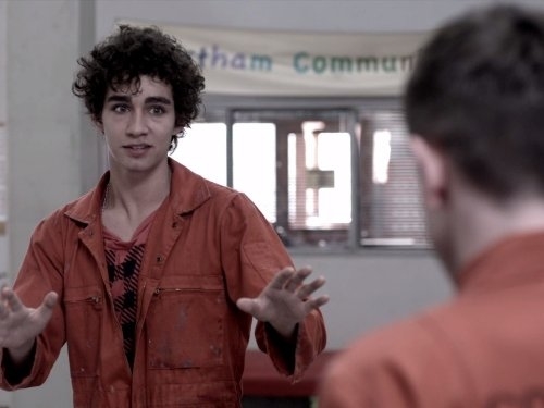 Robert Sheehan, wearing an orange jacket, gestures with both hands while talking to an unseen person in a room with a &quot;tham Community&quot; banner