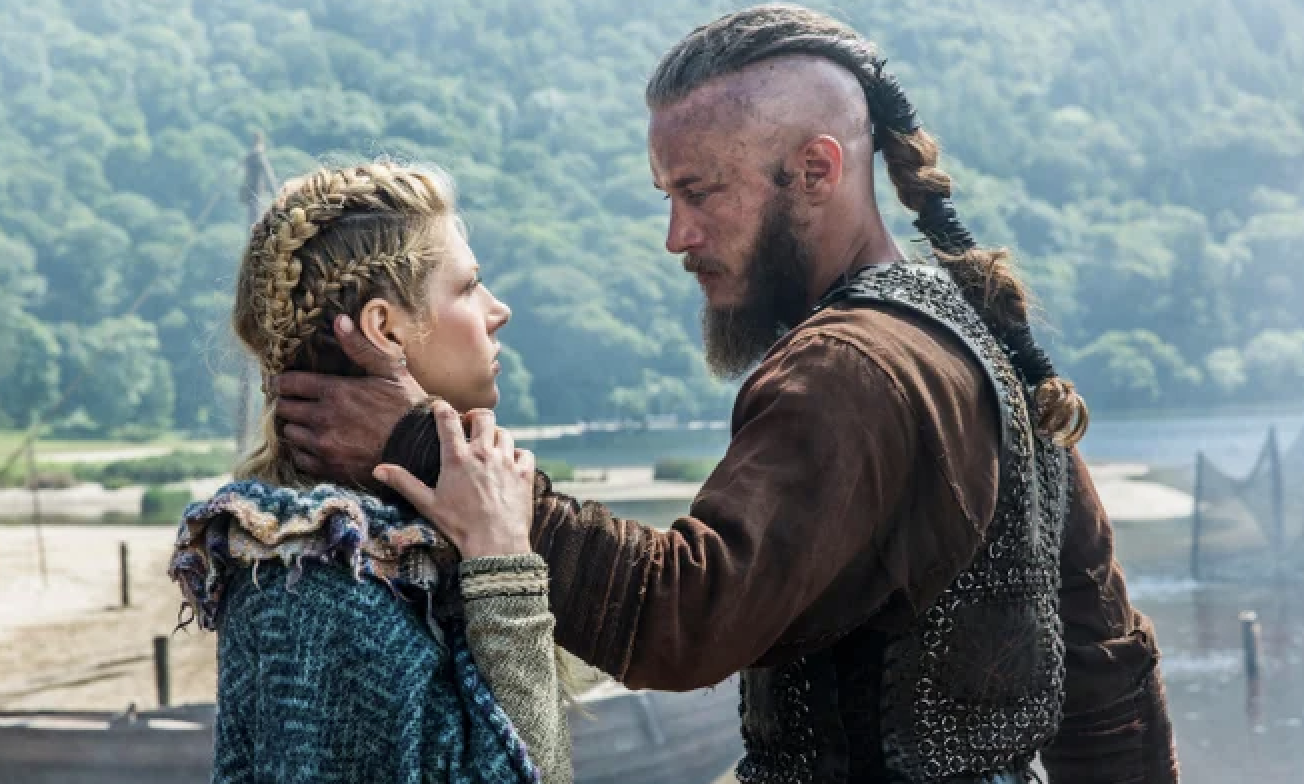 Katheryn Winnick and Travis Fimmel, in Viking-era costumes, face each other closely with serious expressions in a forested riverside setting