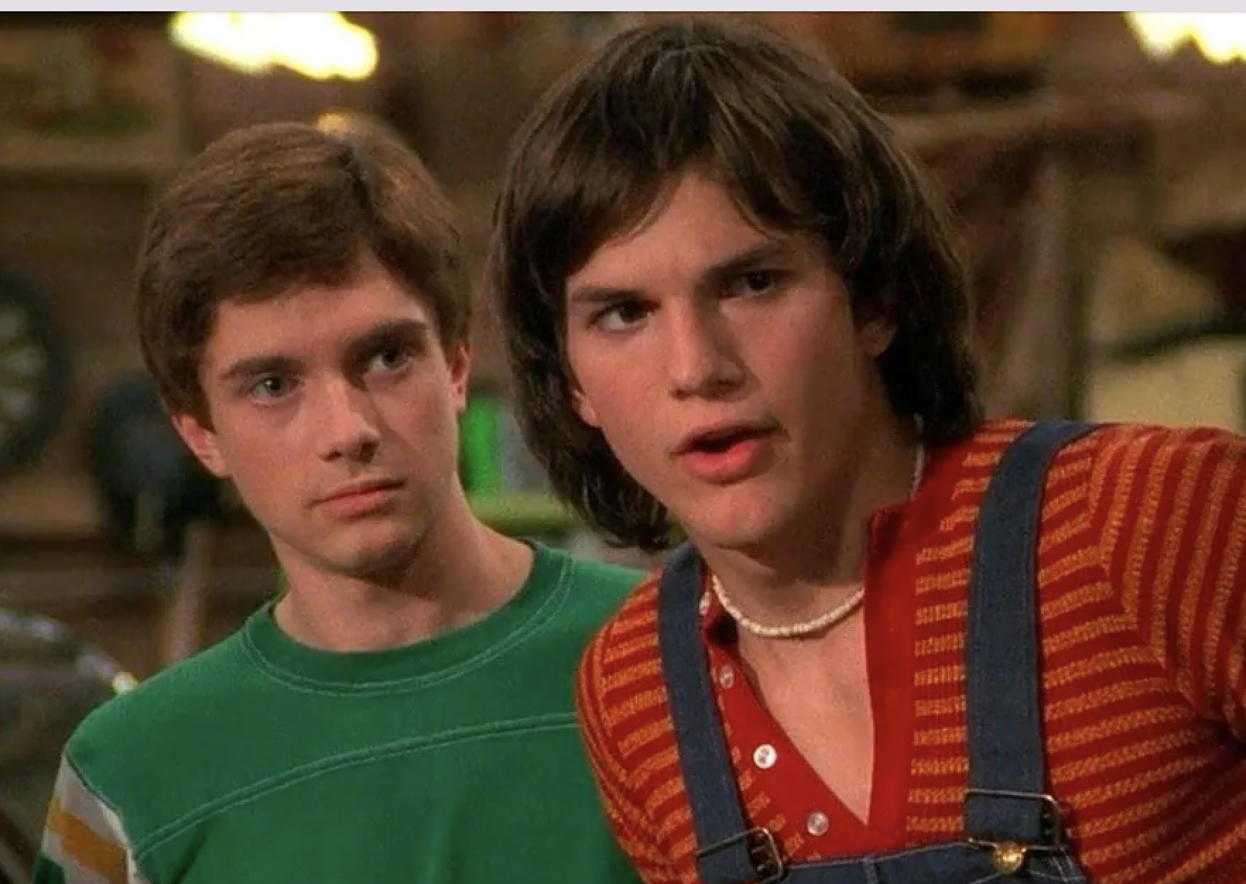Topher Grace and Ashton Kutcher in a scene with casual 1970s attire. Ashton wears denim overalls over a striped shirt with a shell necklace and Topher in a green sweater