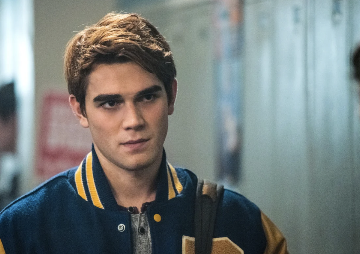 K.J. Apa standing in a school hallway wearing a varsity jacket, gazing intently ahead with his satchel strap over his shoulder