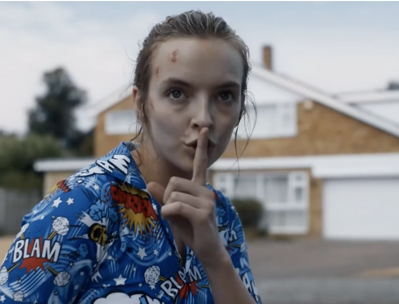 Jodie Comer stands outside a suburban house making a shushing gesture with her finger to her lips, dressed in a comic-themed onesie