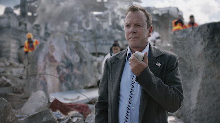 Kiefer Sutherland stands in a suit holding a radio in a disaster zone with workers in the background