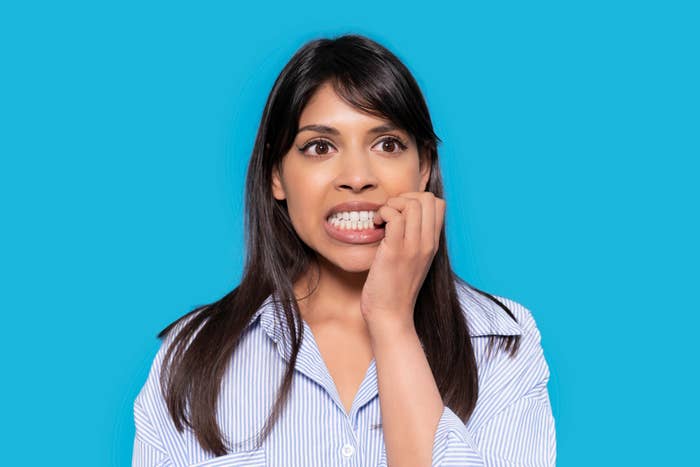 A woman with long dark hair in a striped shirt is anxiously biting her nails against a plain background