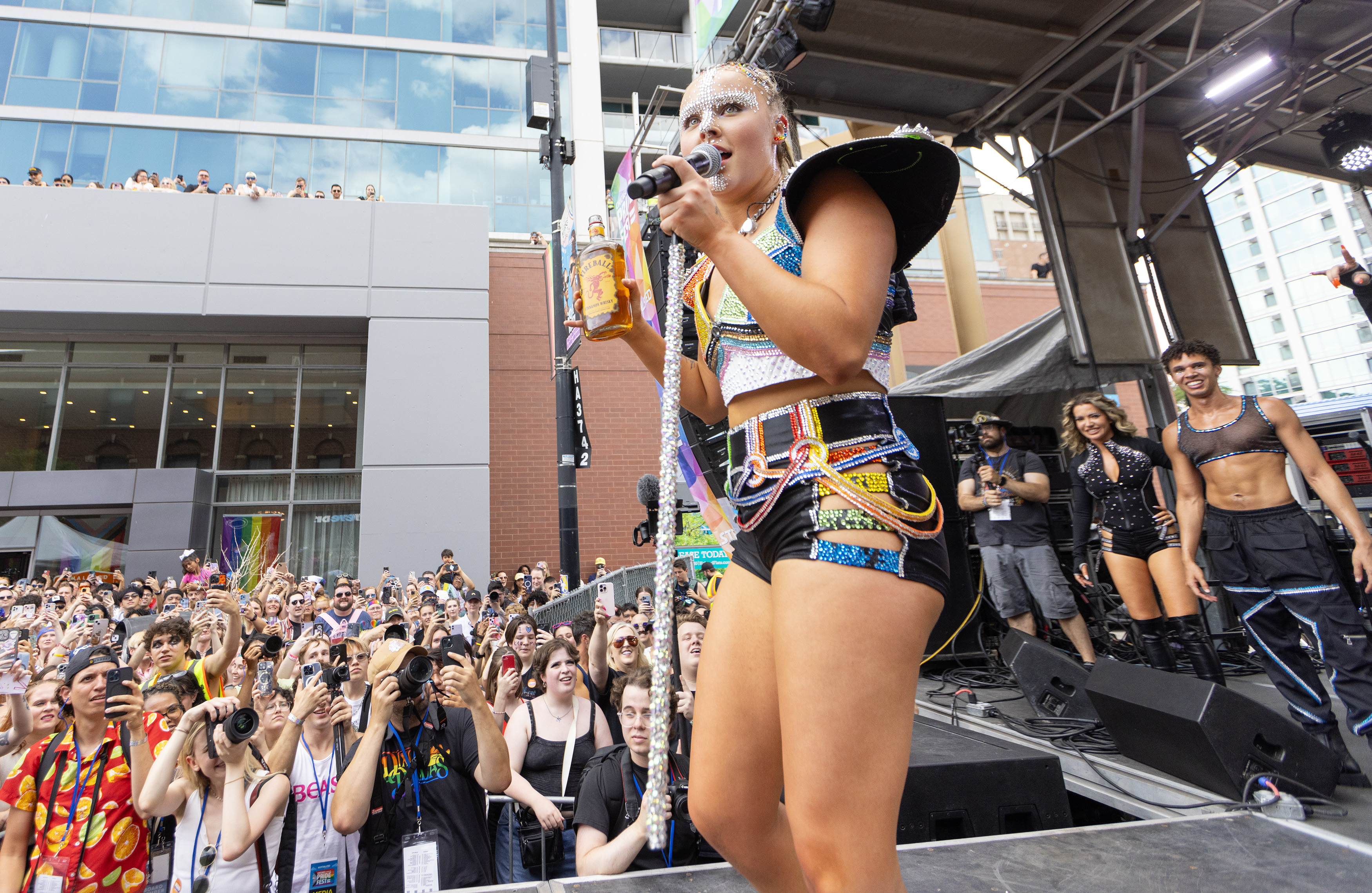 JoJo Siwa performs at an outdoor concert in a futuristic stage outfit with a mic in one hand and a bottle in the other. The crowd cheers and takes photos