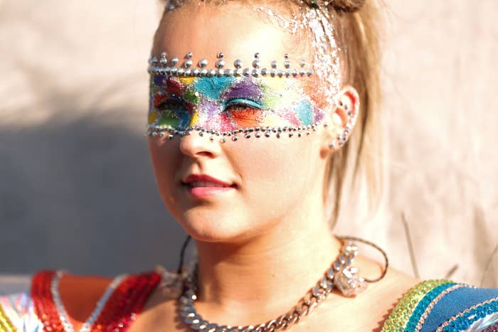 JoJo Siwa wearing a colorful, gem-studded eye mask and a vibrant outfit with a chain necklace, posing outdoors