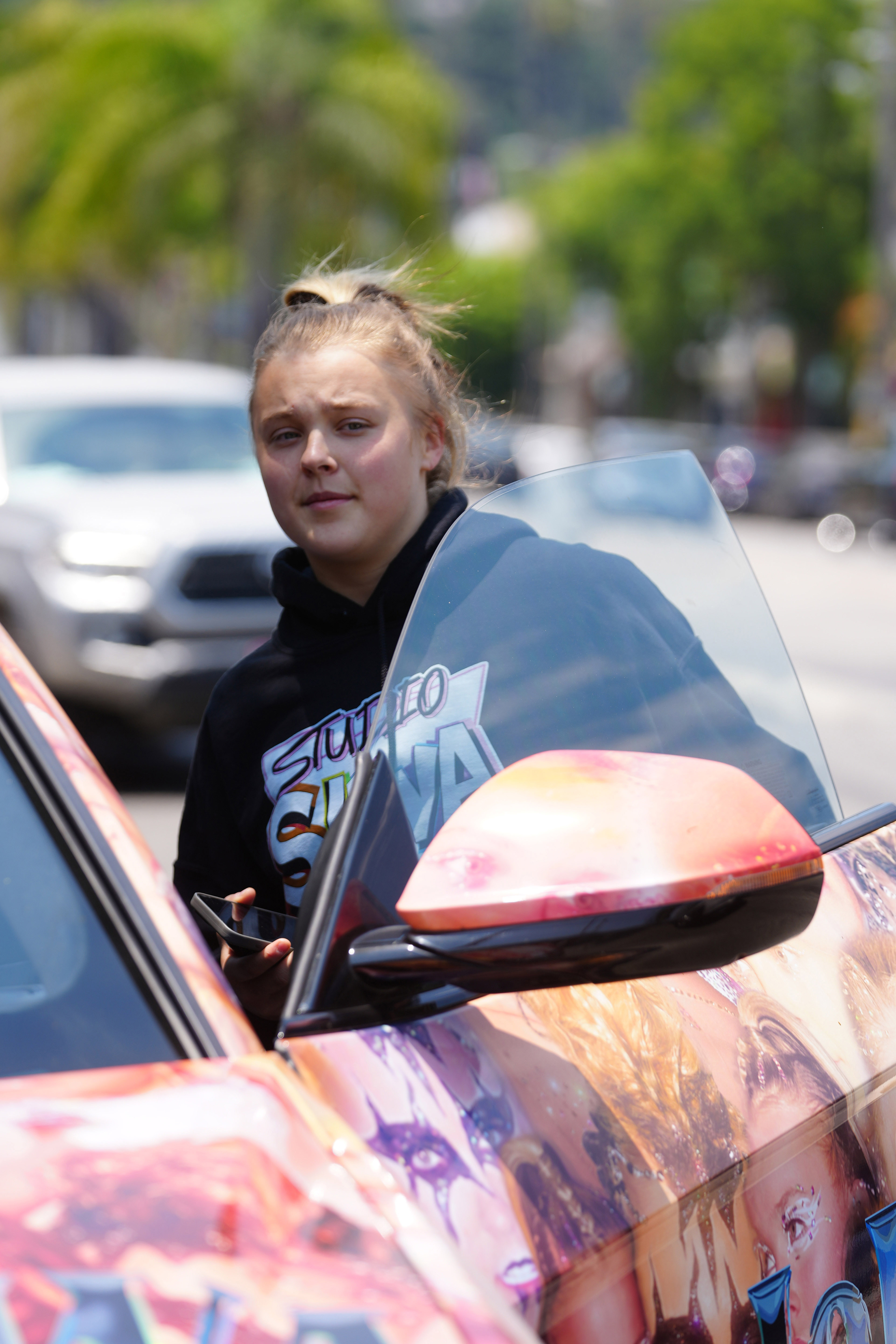 JoJo Siwa getting into a colorful car decorated with artistic designs, holding a phone in one hand while wearing a black hoodie with "Siwa" written on it