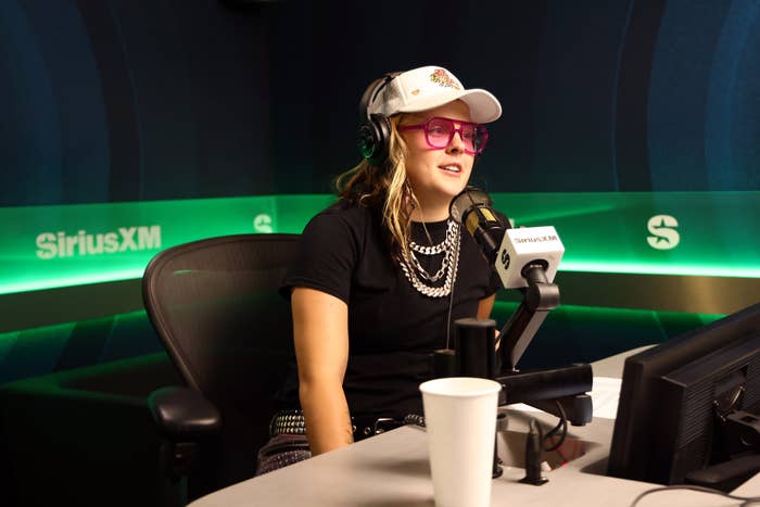 A person in casual attire, wearing a white cap and pink glasses, sits in a SiriusXM studio for an interview