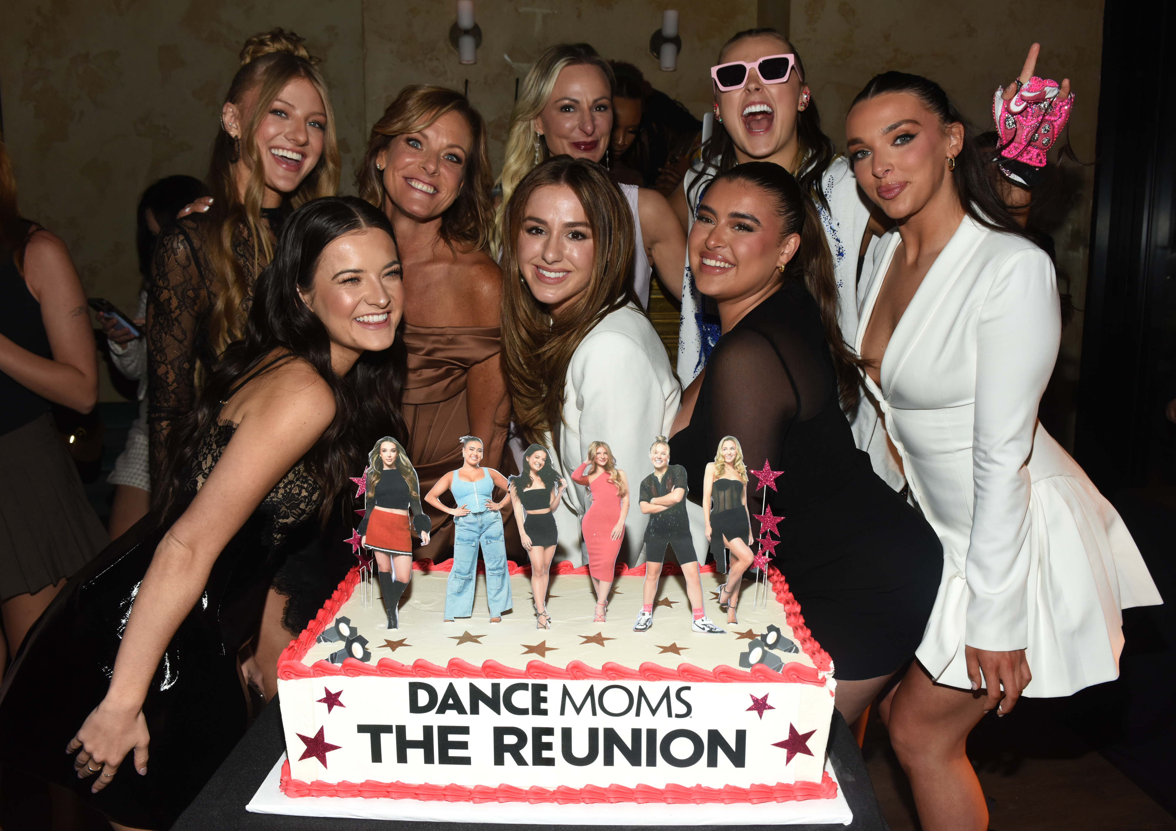 Dance Moms reunion with cast members smiling around a themed cake that reads "Dance Moms The Reunion."