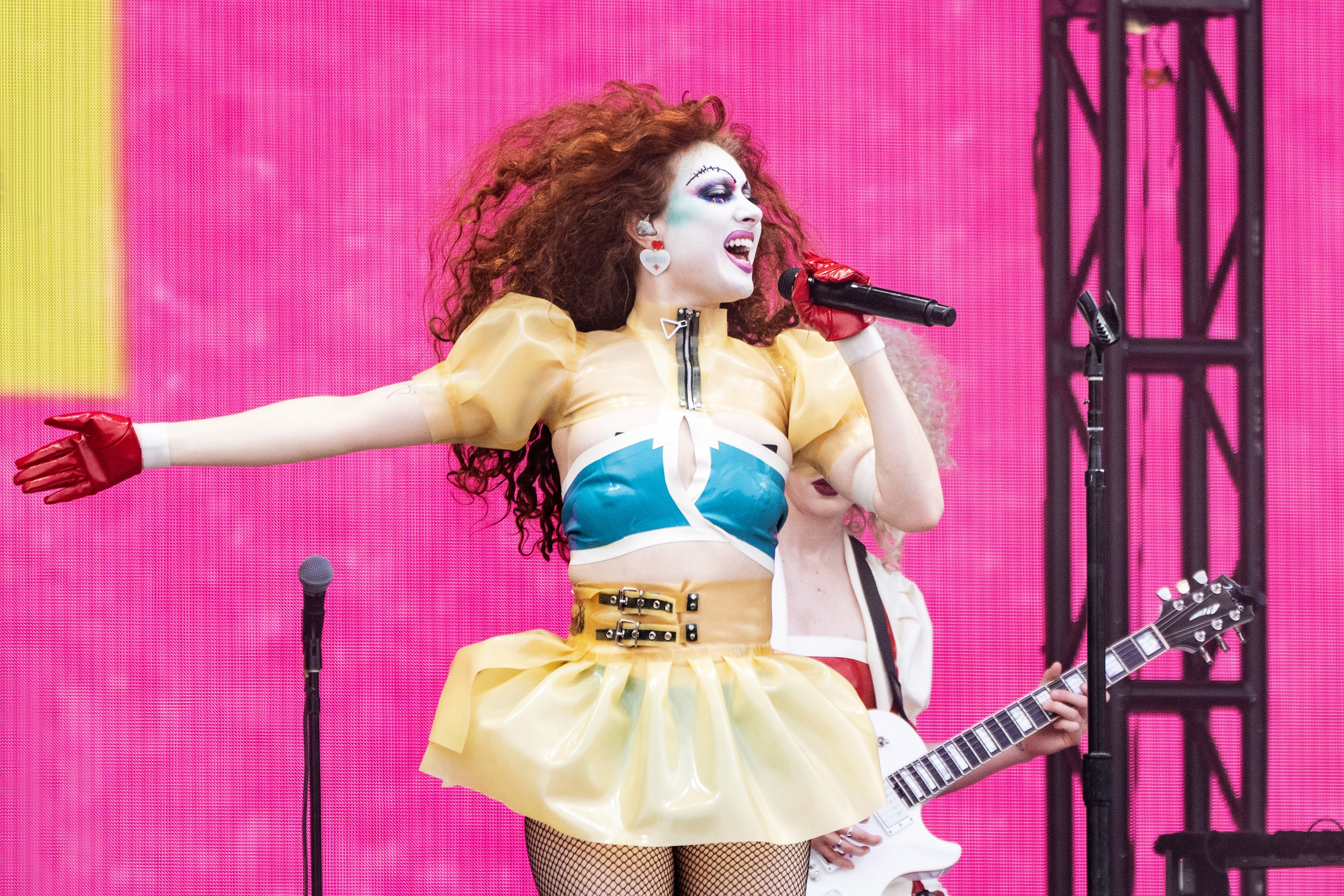 Performer in bold stage makeup and a vibrant outfit with a blue top and yellow skirt sings passionately on stage. Guitarist plays in the background