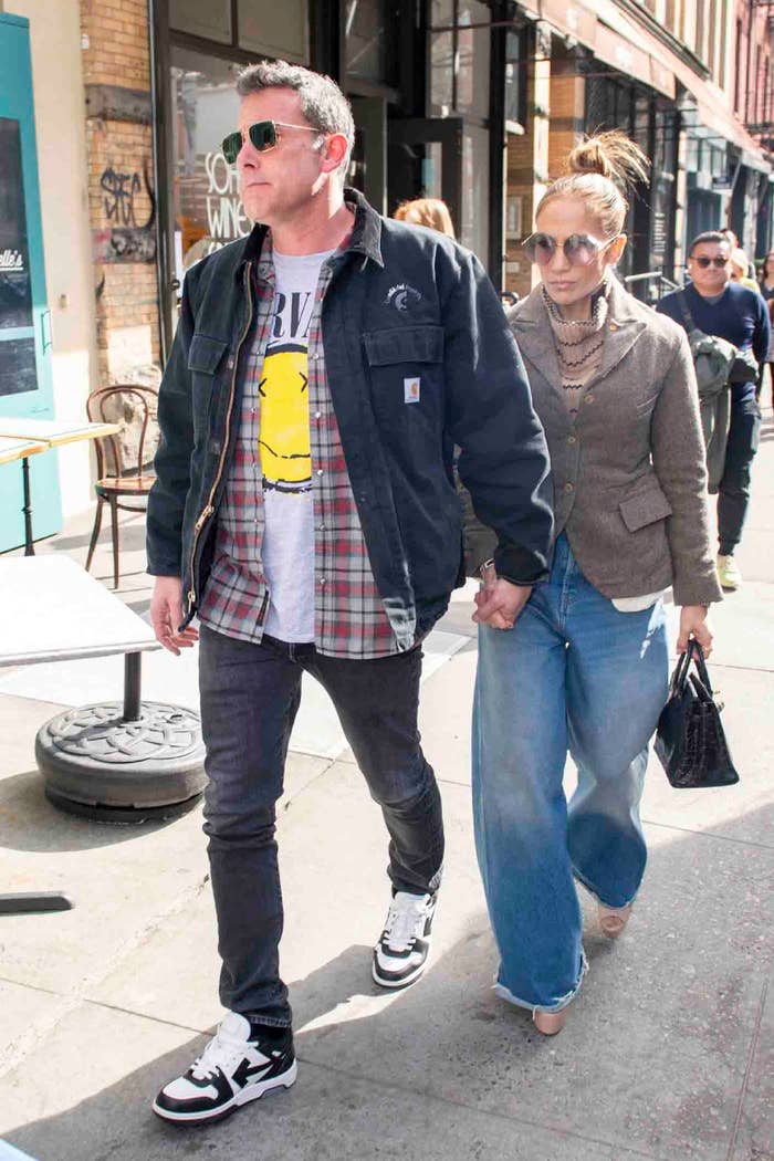 Ben Affleck and Jennifer Lopez hold hands while walking. Ben wears casual attire with a T-shirt, plaid shirt, and jacket. Jennifer wears a blazer and wide-leg jeans