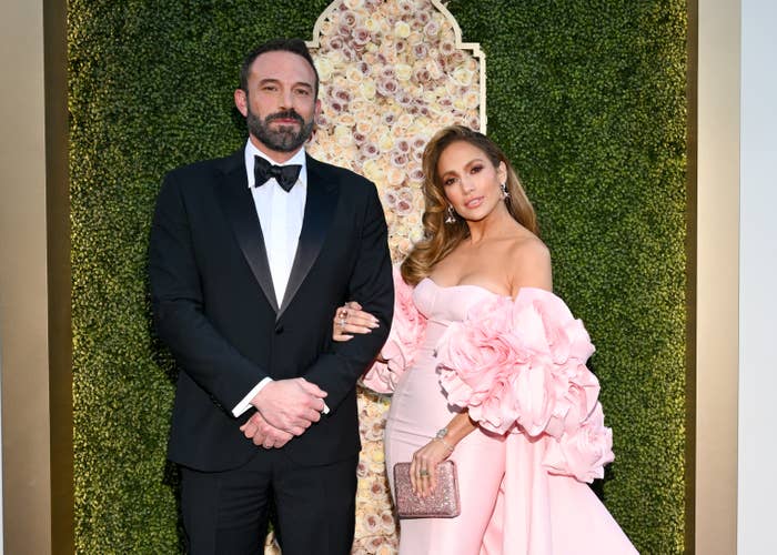 Ben Affleck and Jennifer Lopez at an event. Ben is in a black tuxedo, and Jennifer is in an elegant off-shoulder gown with floral embellishments