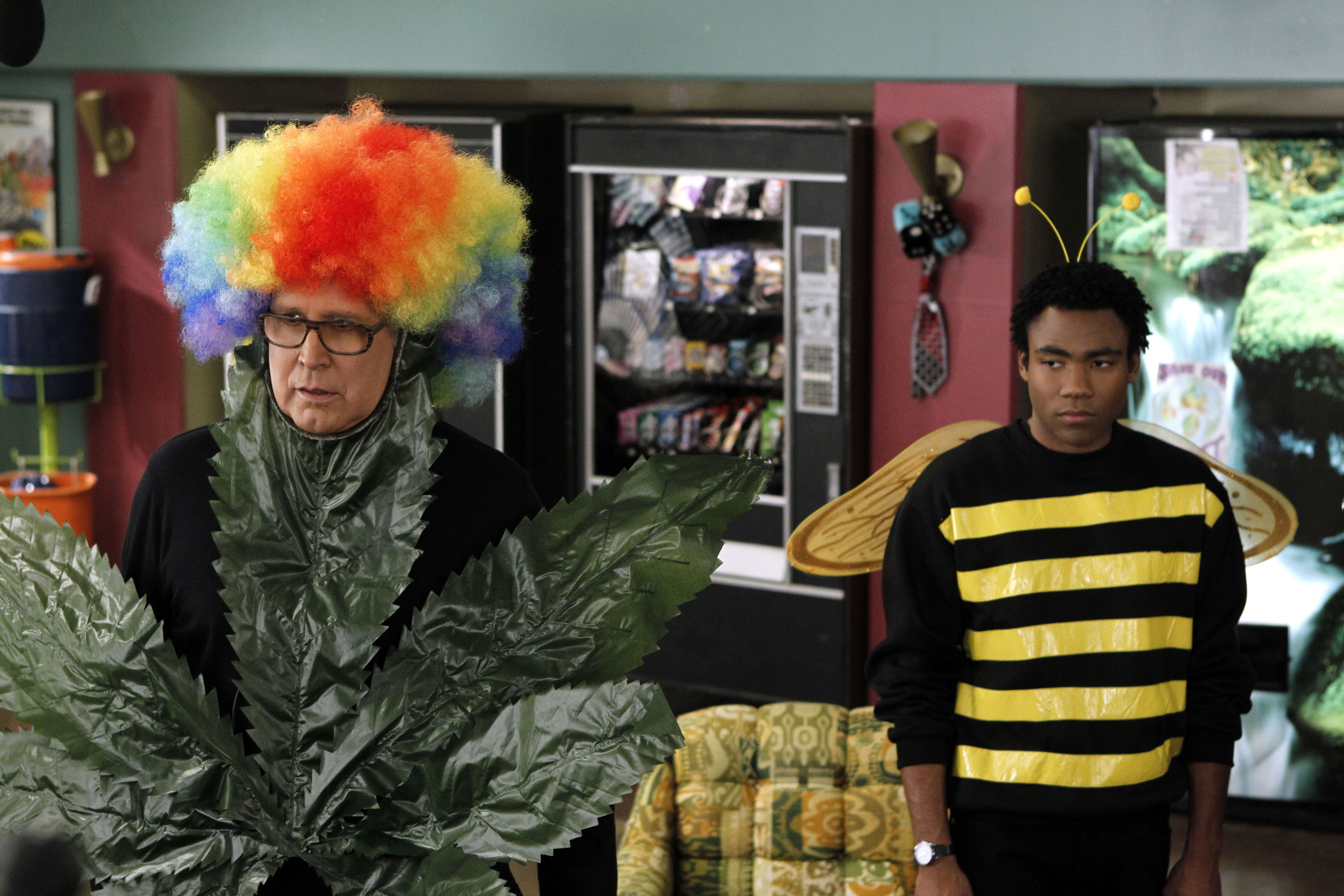 Chevy Chase dressed as a giant cannabis leaf and Donald Glover dressed as a bee, both standing indoors with vending machines in the background