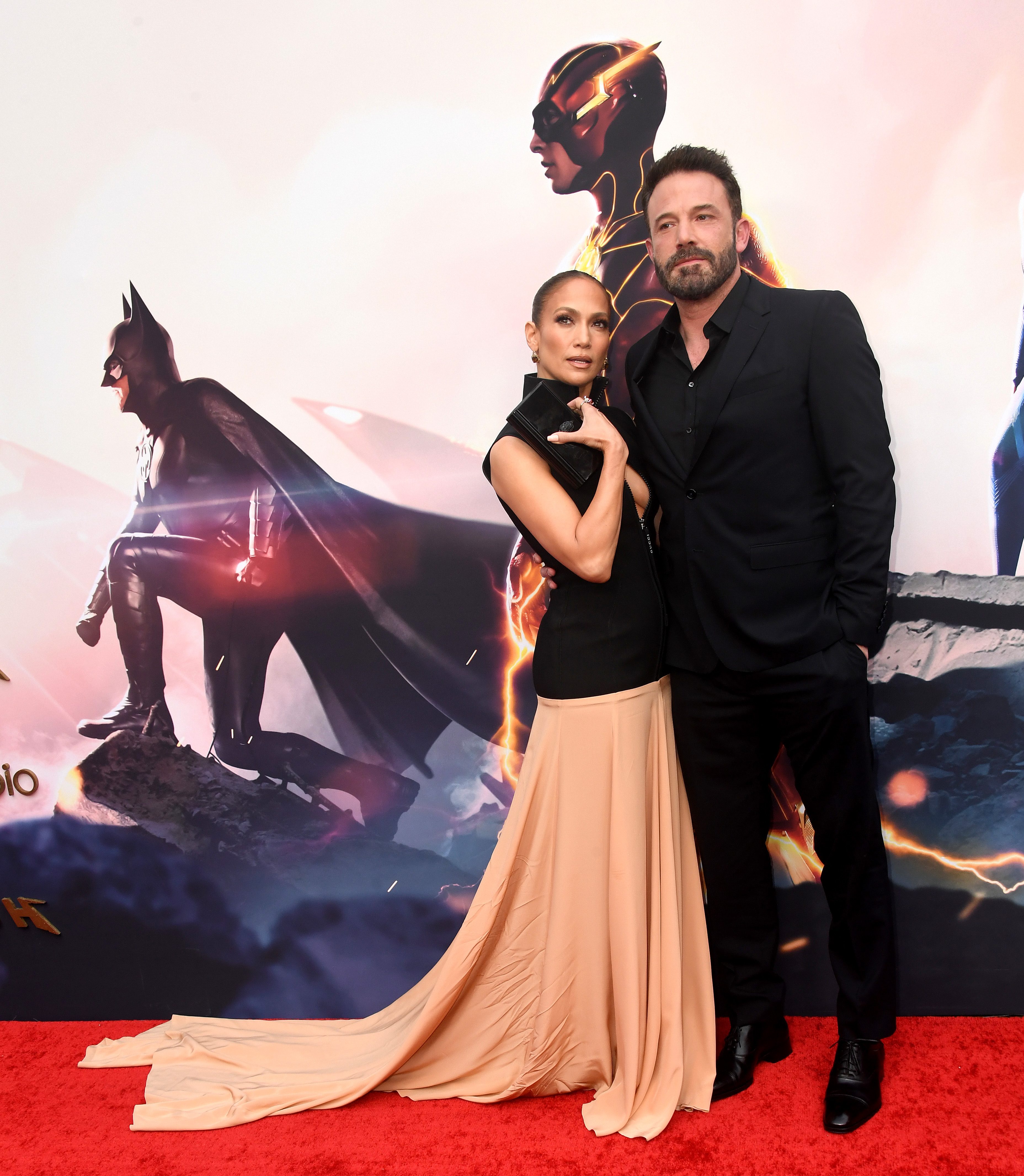 Jennifer Lopez in a black and beige gown and Ben Affleck in a black suit pose together on a red carpet event with superhero posters in the background