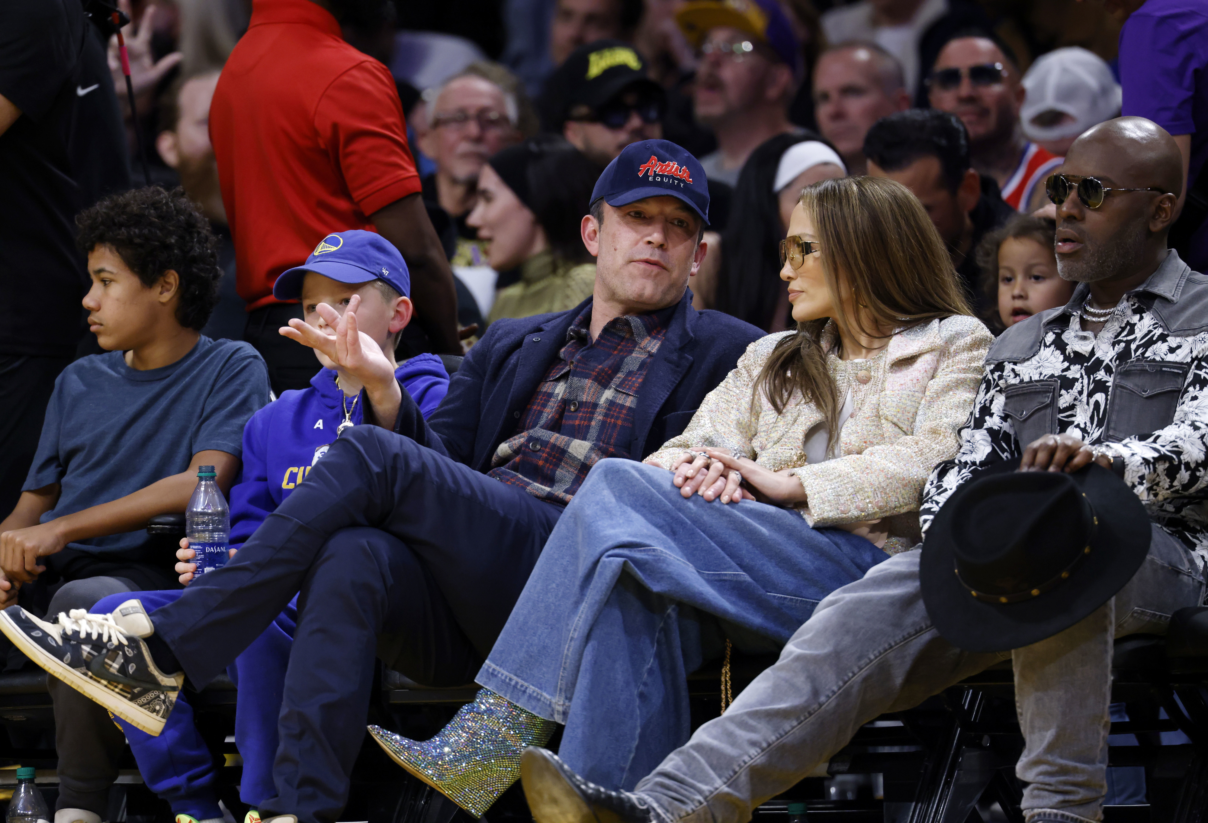 Ben Affleck, sitting with Jennifer Lopez, wearing casual clothes at a basketball game. Others sit nearby, including a child in a blue cap and a man in a black hat
