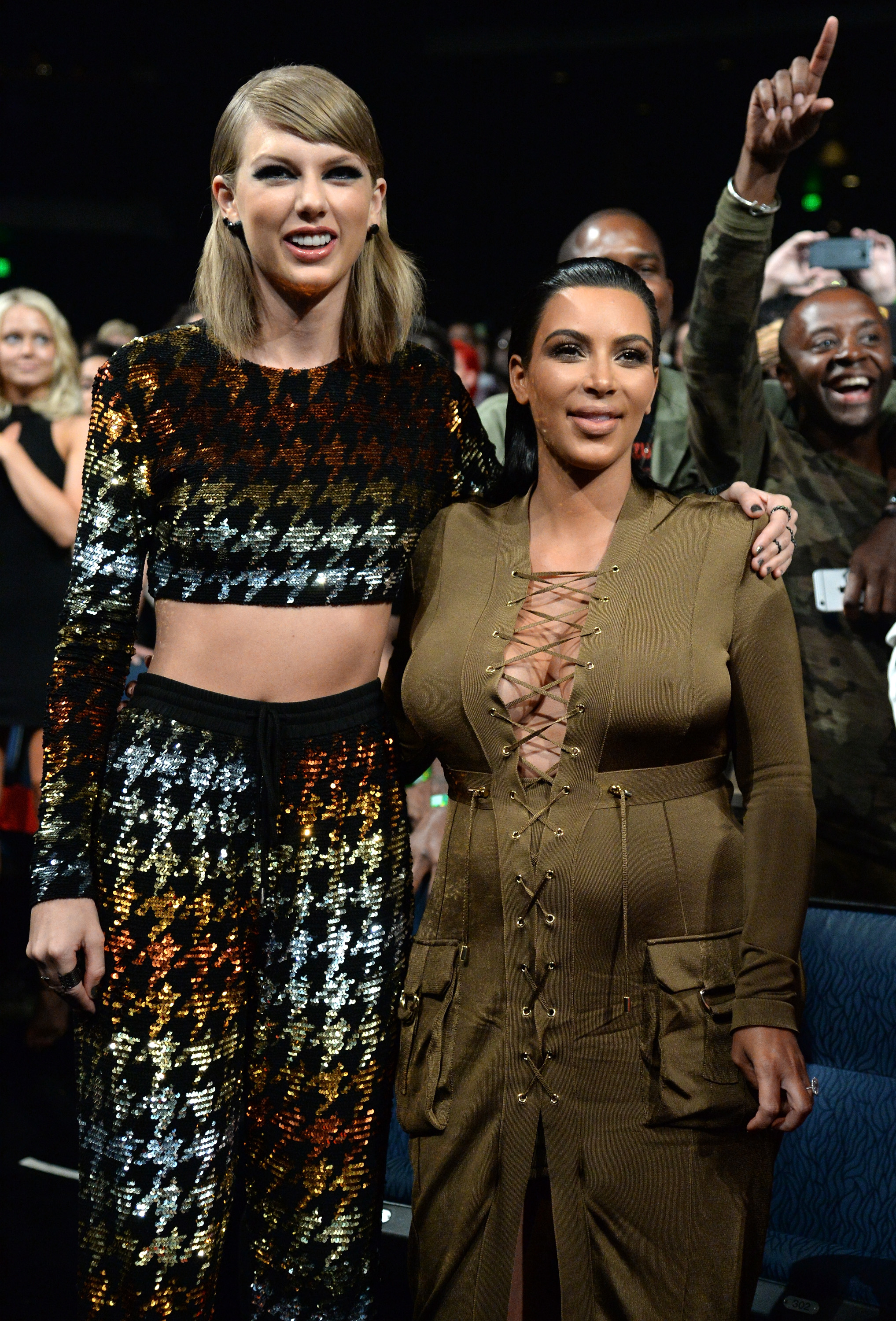 Taylor Swift in a sequined crop top and pants, and Kim Kardashian in a lace-up dress, at a public event with people in the background