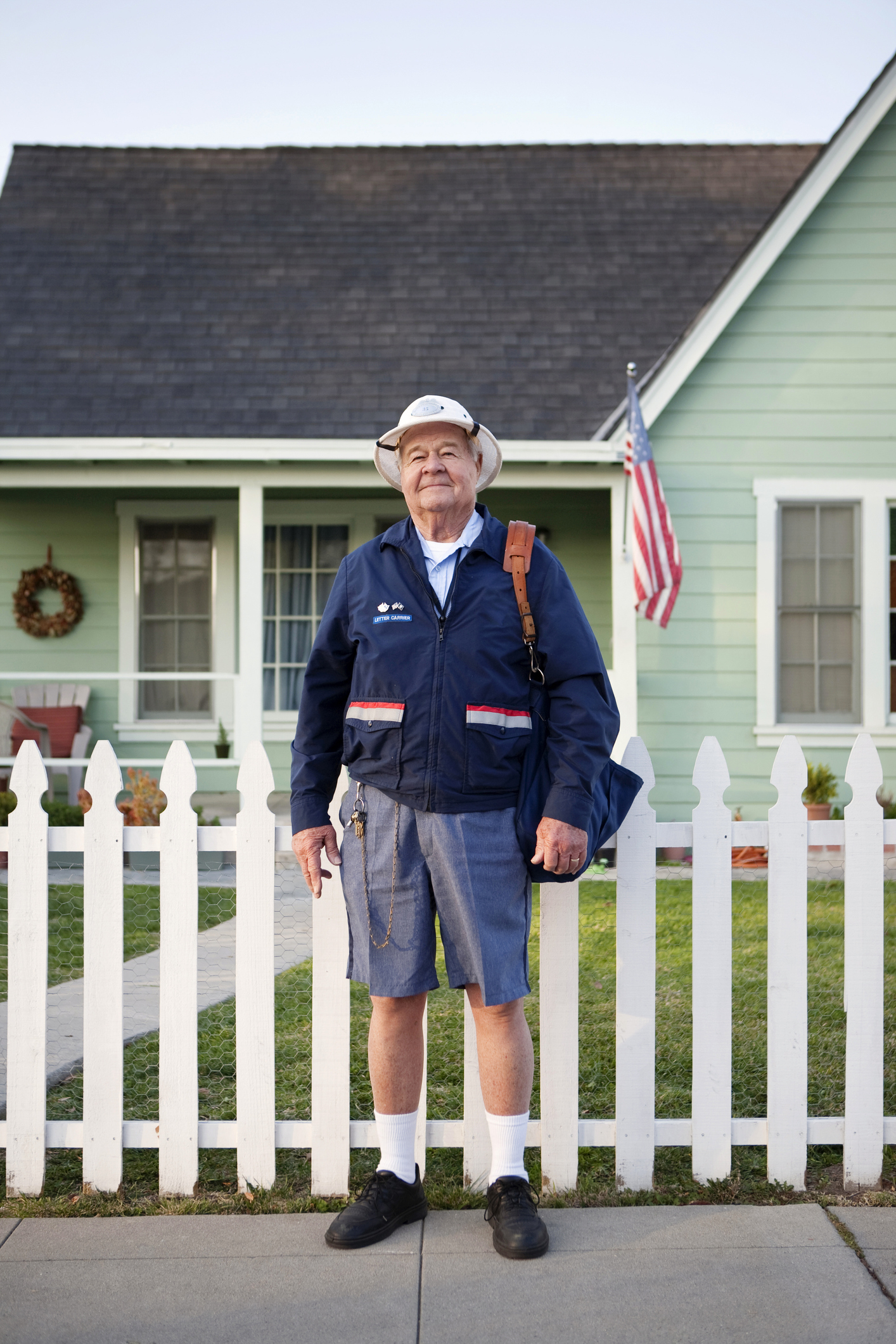 A senior mail carrier wearing a uniform, including shorts and a hat, stands in front of a white picket fence and a house with an American flag