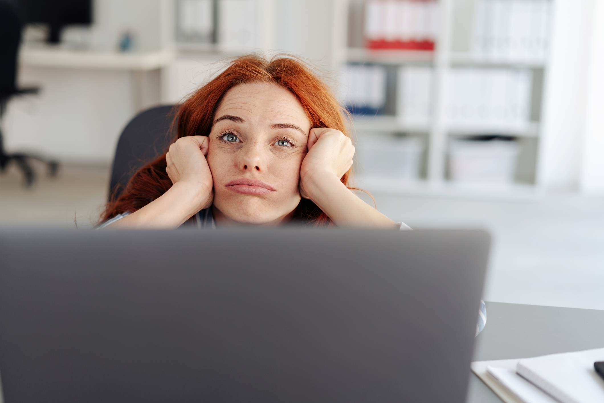 A person with long, straight hair rests their head in their hands while looking at a laptop in an office. Shelves with binders are visible in the background