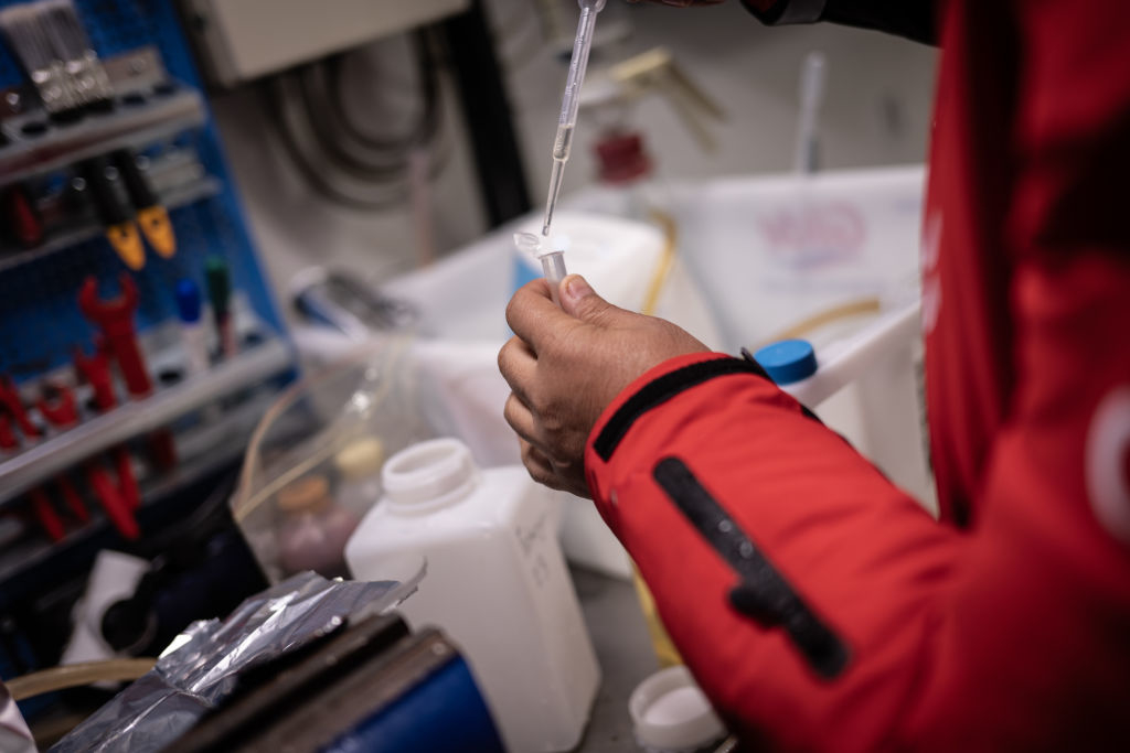 Person in a work setting using a pipette to dispense liquid into a small container, with various lab equipment and bottles visible in the background