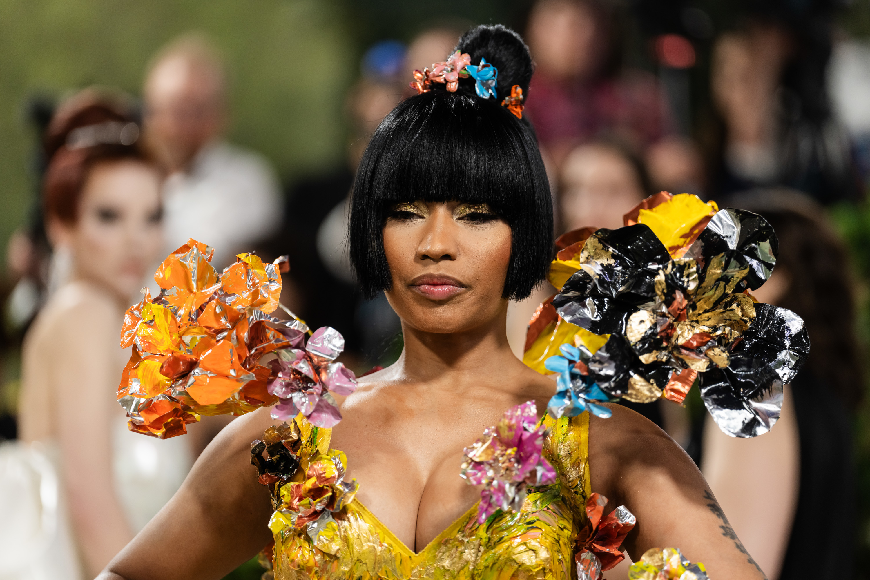 Nicki Minaj at a red carpet event, wearing a bold, floral-themed gown with vibrant, sculpted flower decorations on her shoulders and chest