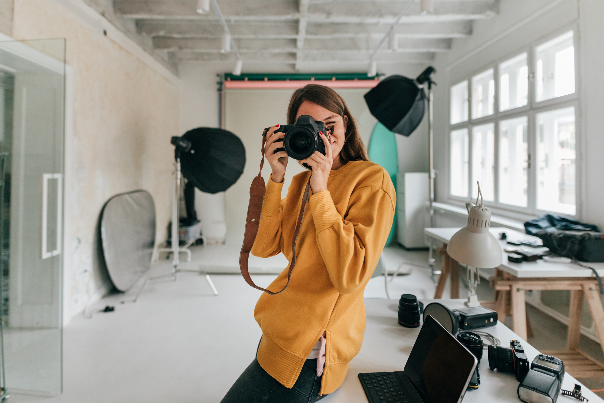 Person in a casual outfit holds a camera up to their face in a professional photography studio filled with equipment