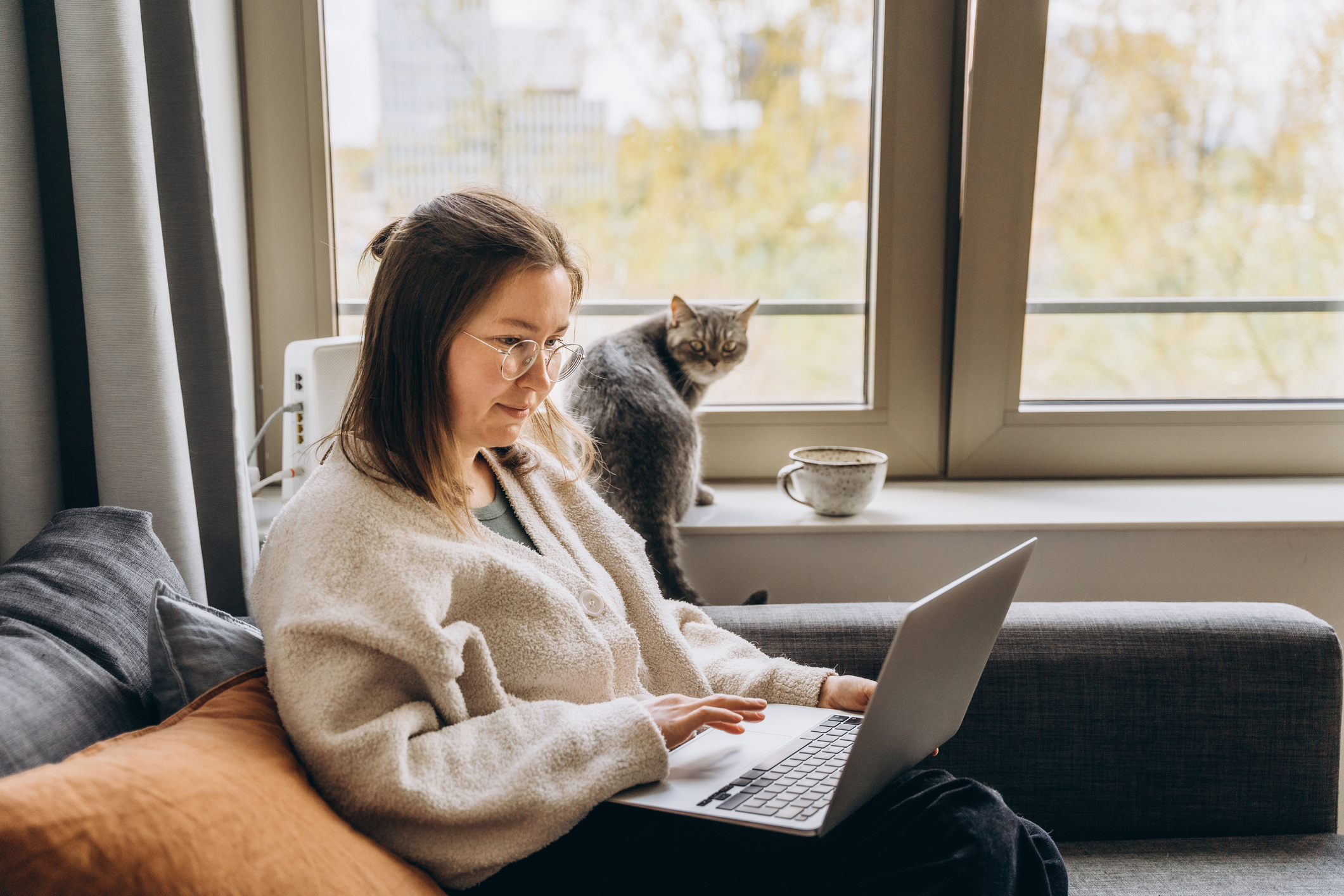 A woman works on a laptop while sitting on a couch with a cat looking out of the window beside her in a cozy home setting