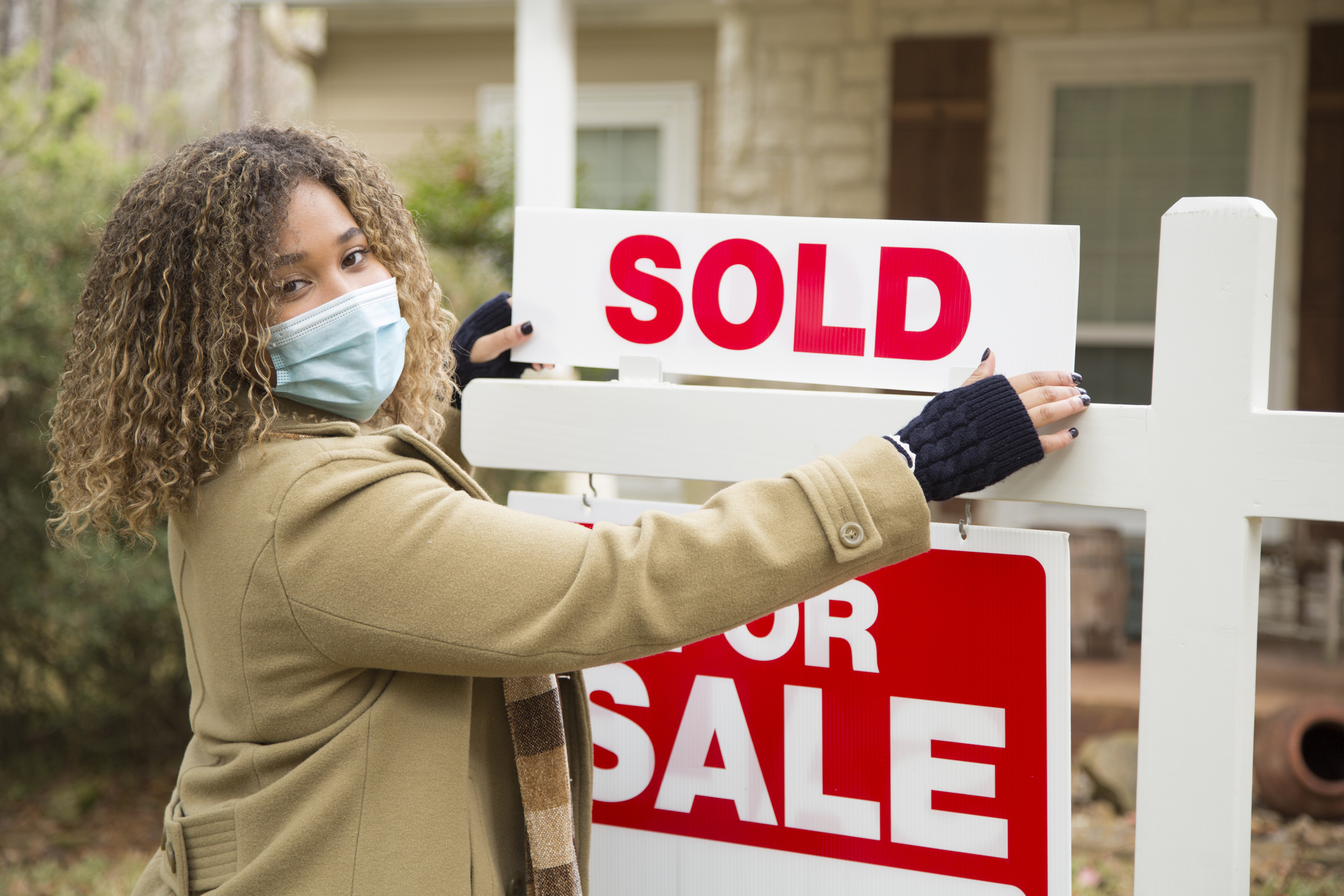 A person wearing a mask, holding a "SOLD" sign over a "FOR SALE" sign in front of a house