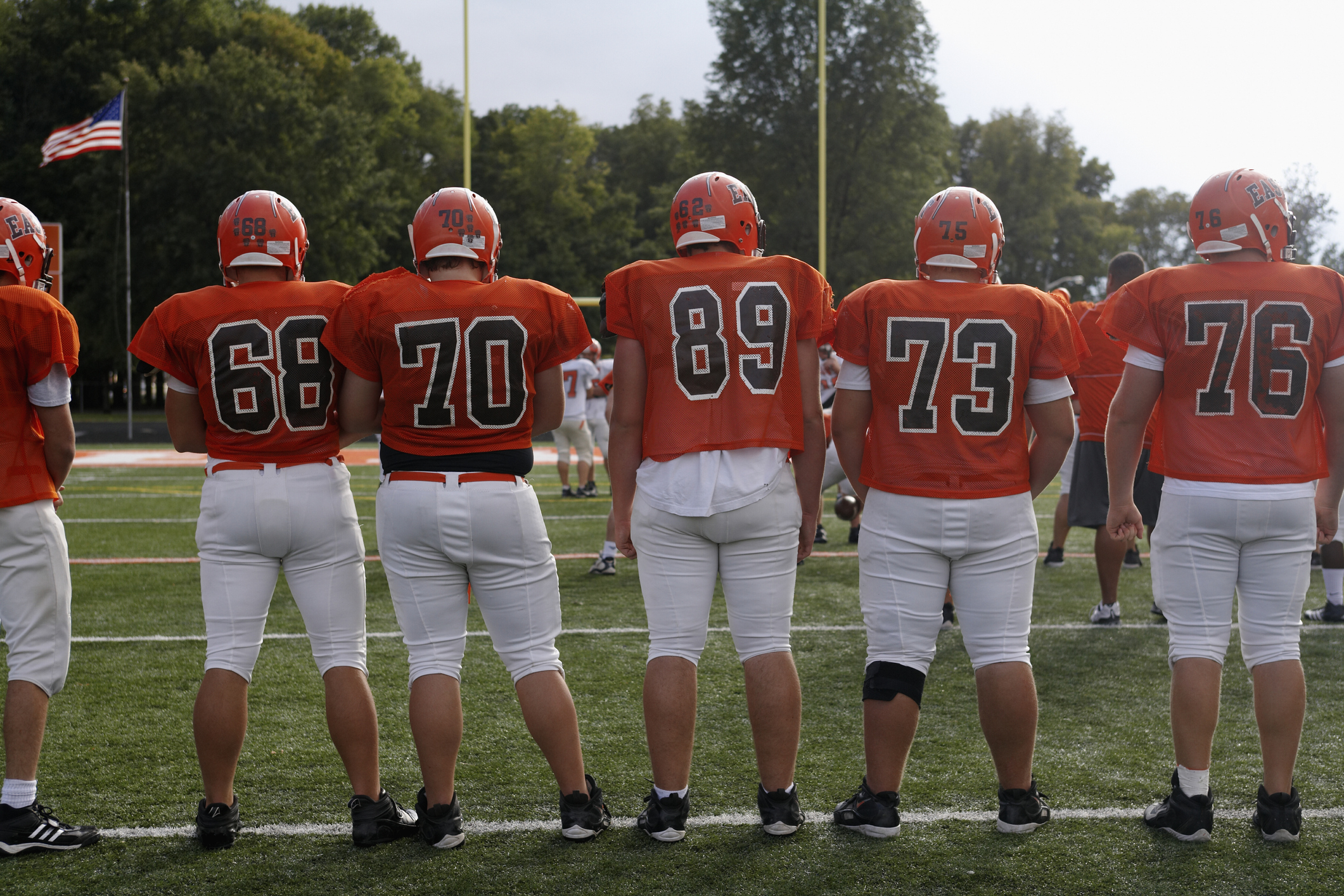 Five football players in uniform stand on the field with numbers 68, 70, 89, 73, and 76 on their jerseys, facing away from the camera. The image is related to Work & Money