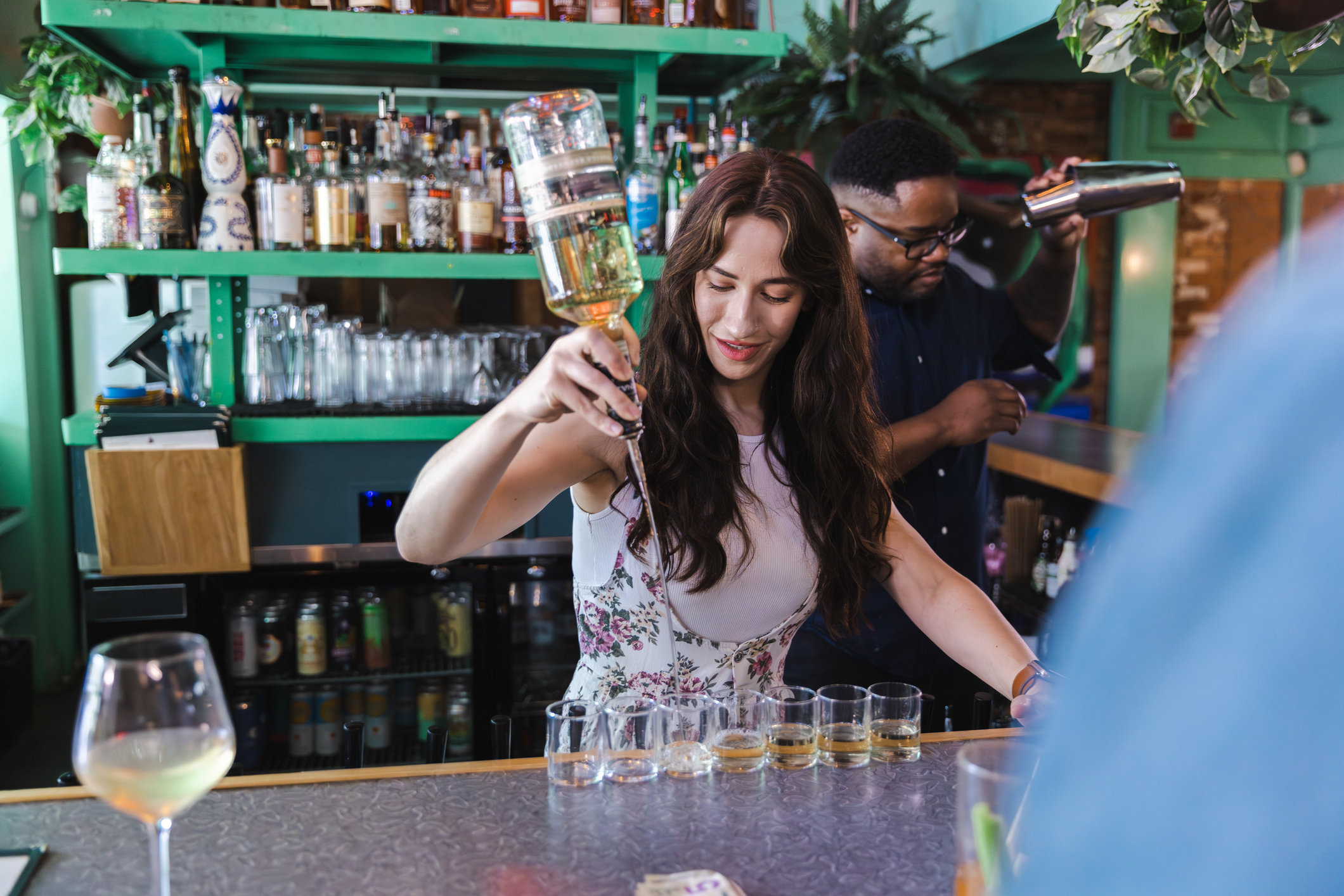 Woman, Ciara Gordon, pours drinks behind a bar while a bartender works in the background. Shelves of bottles and various bar tools are visible