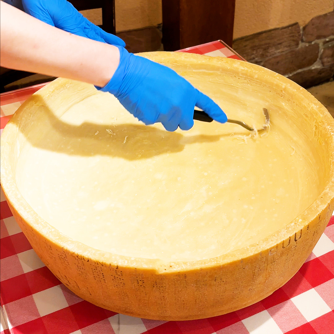 大きなチーズの器の中を青い手袋をした手が調理器具で混ぜている様子。